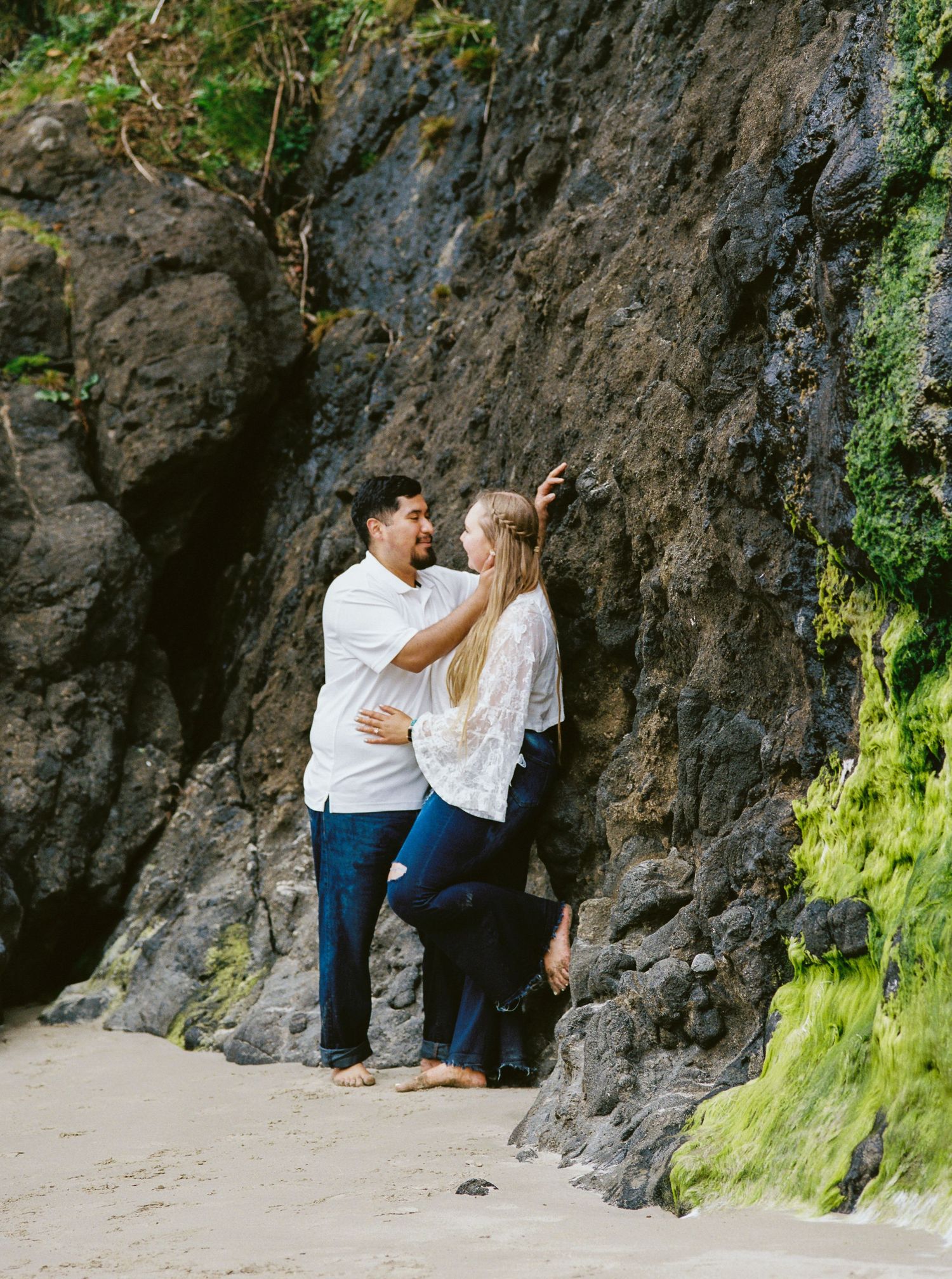 Couple sharing tender moment against mossy coastal rock formation on Oregon beach.