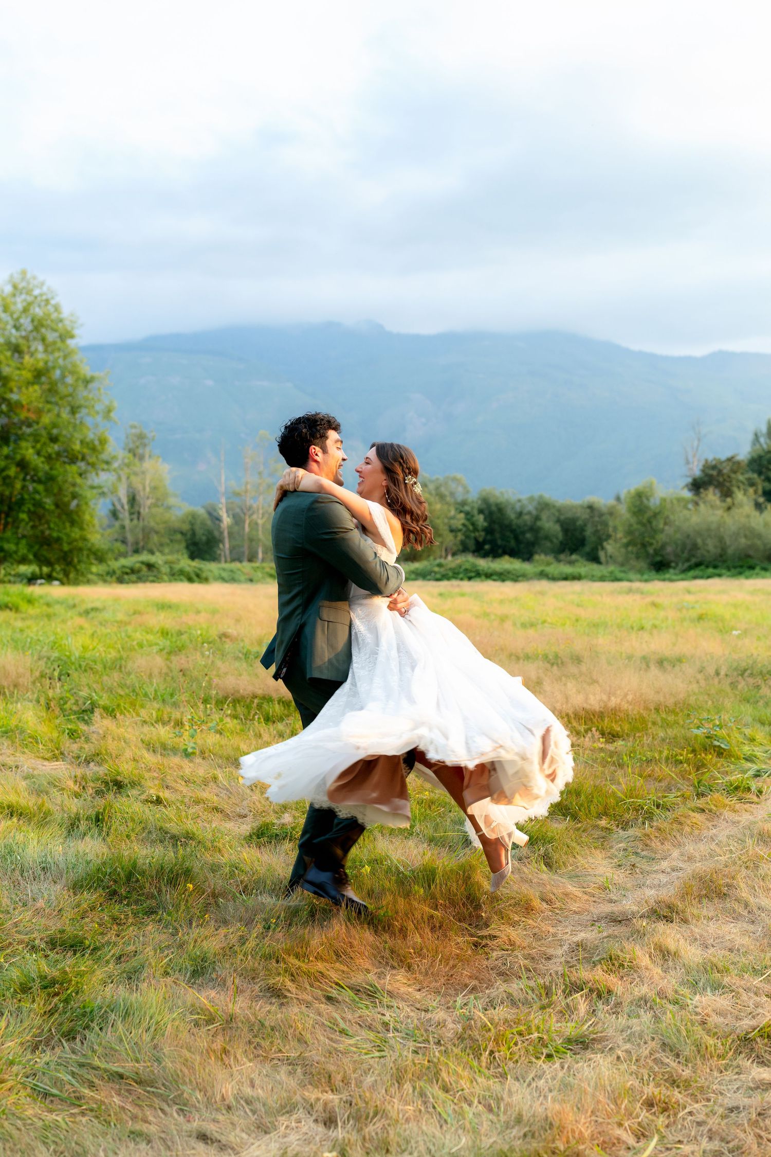 Groom lifting bride in white dress, spinning in a mountain meadow on their wedding day.