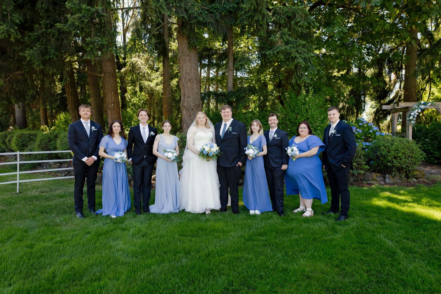 Wedding party poses outdoors with bride and groom flanked by bridesmaids in blue dresses and groomsmen in black suits.