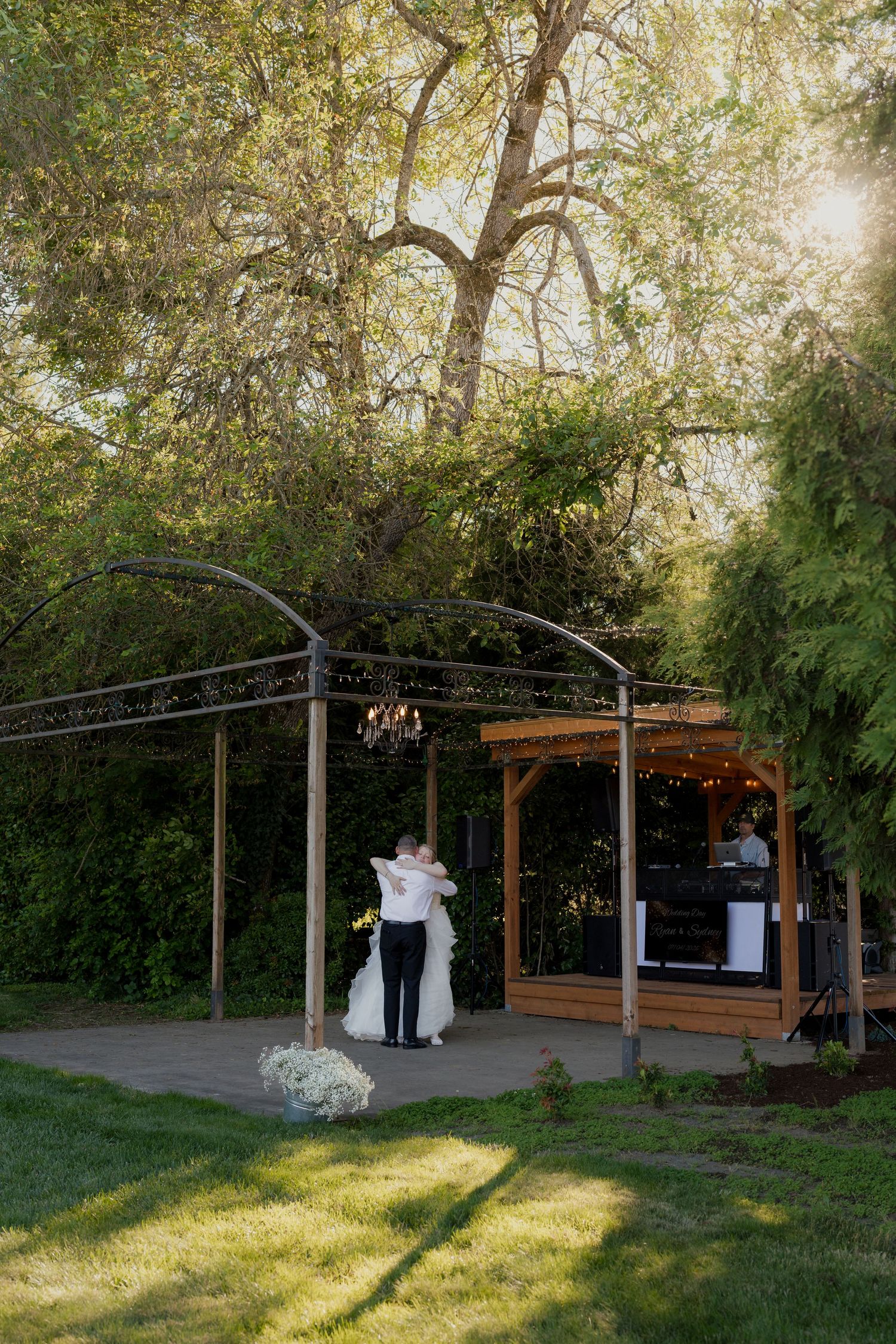 Bride and groom share first dance under an arched pergola with chandelier, surrounded by lush trees at an outdoor wedding venue.