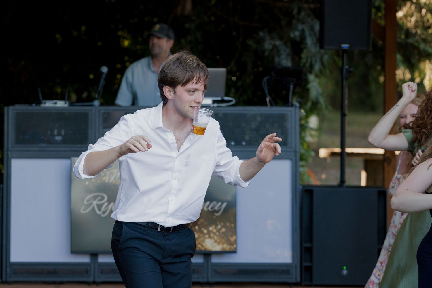 Energetic wedding guest dances with beer glass at outdoor reception near band stage.