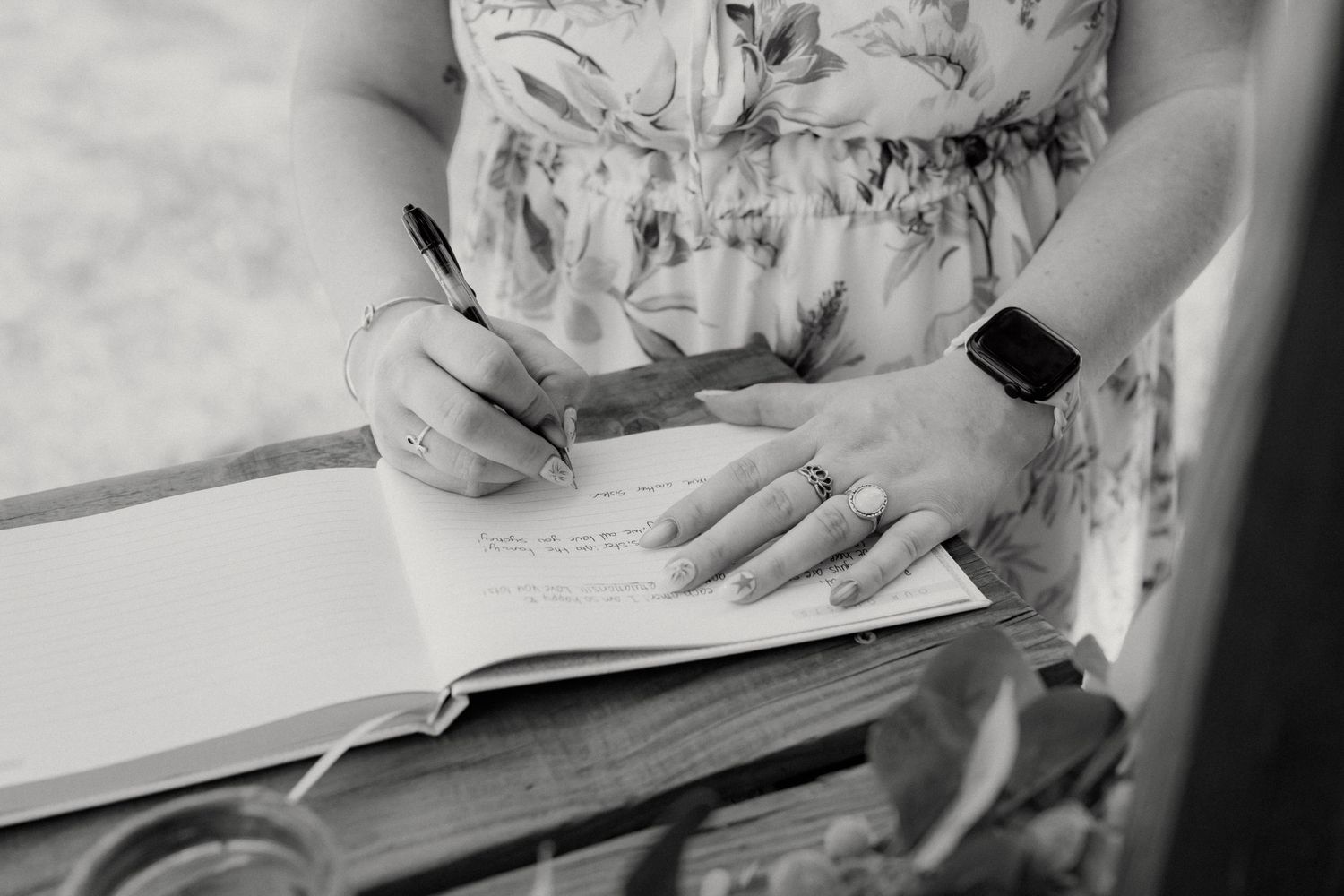 Woman in floral dress signing a guest book with a pen, wearing a smartwatch and ring, black and white photo.