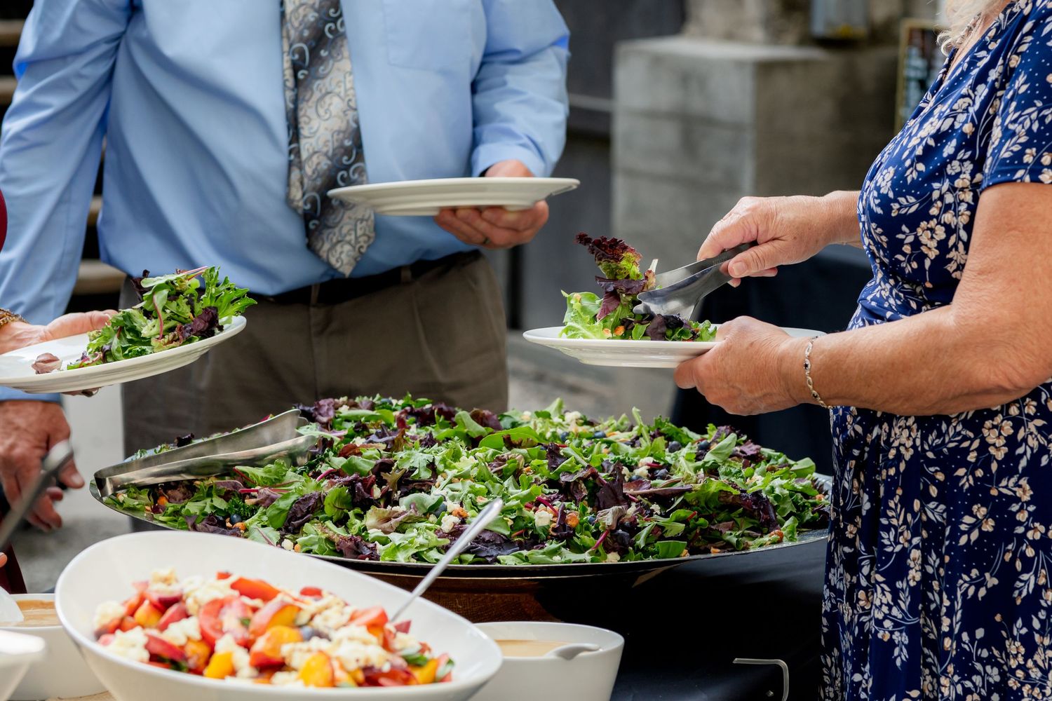 Wedding guests serving themselves fresh green salad and colorful dishes at an outdoor buffet.
