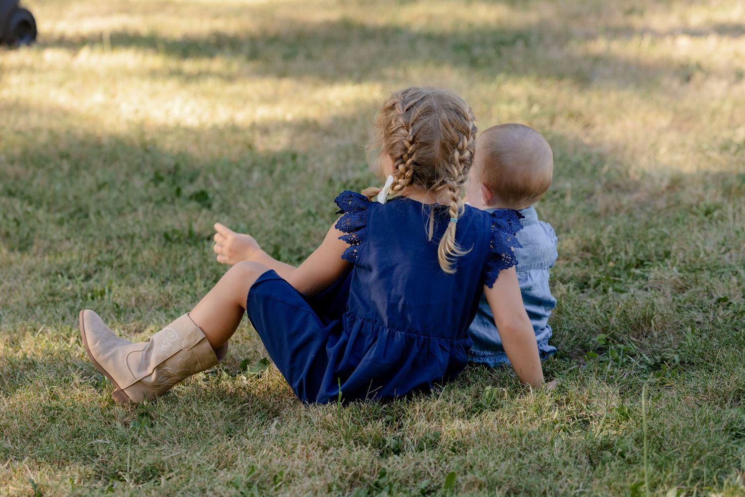 Young girl in navy dress and toddler sitting together on grass at outdoor wedding.