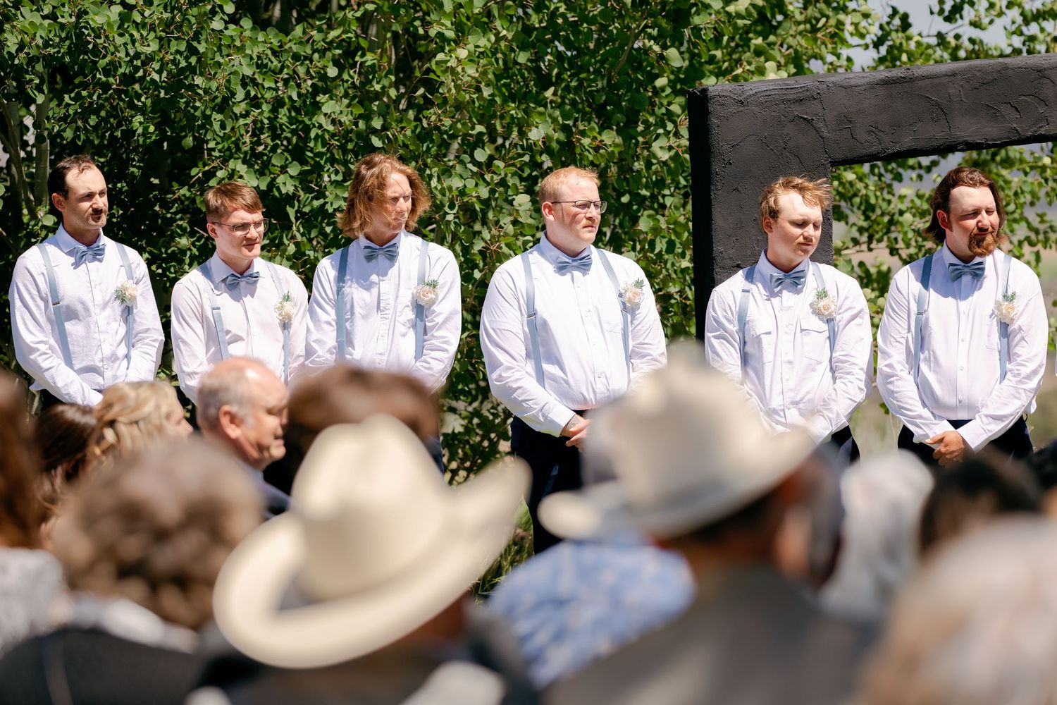 Groomsmen in white shirts and bow ties stand in a line at an outdoor wedding ceremony with guests in foreground.