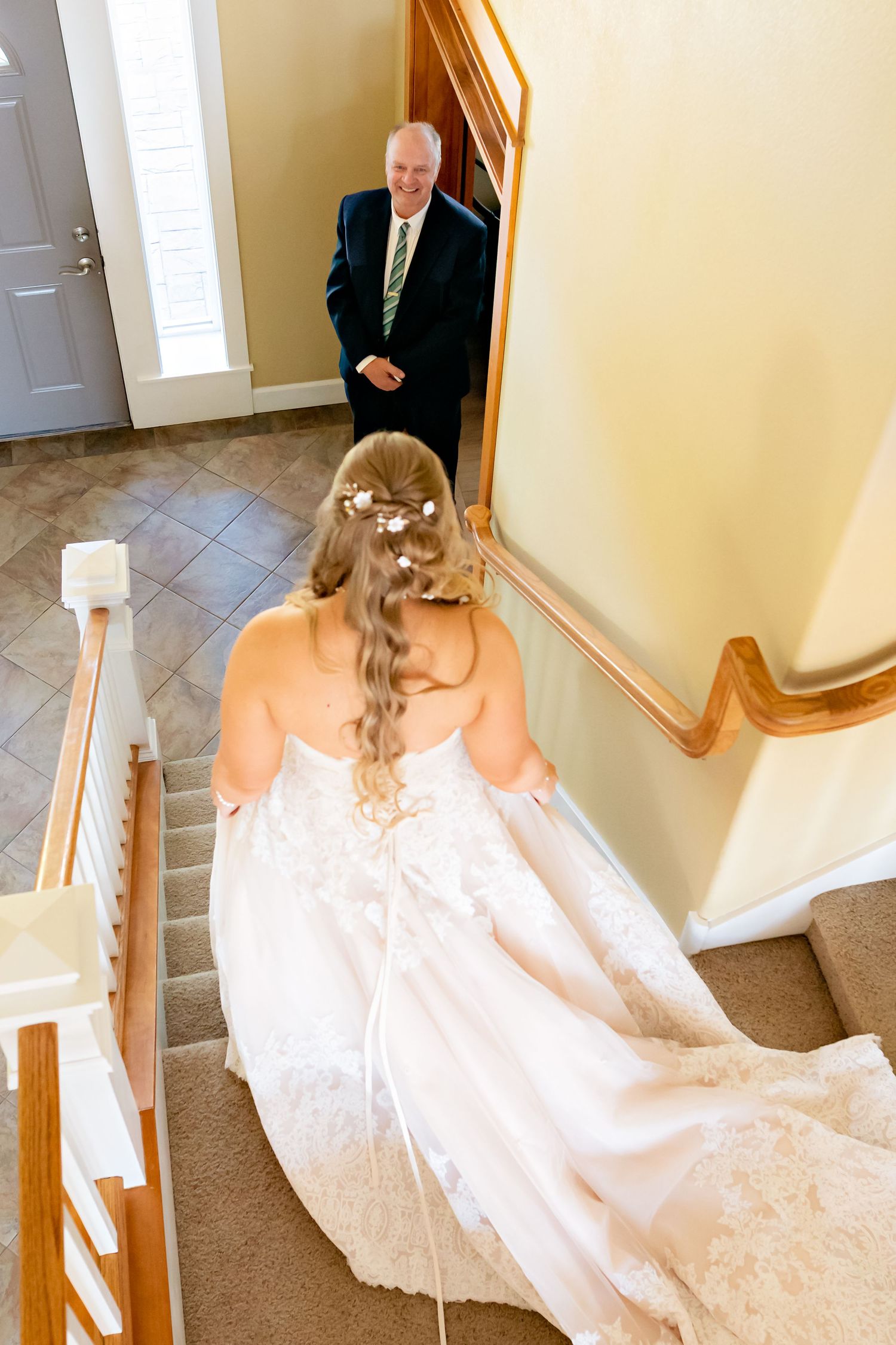 Bride in lace wedding gown descends staircase as emotional father sees her for the first time on wedding day.