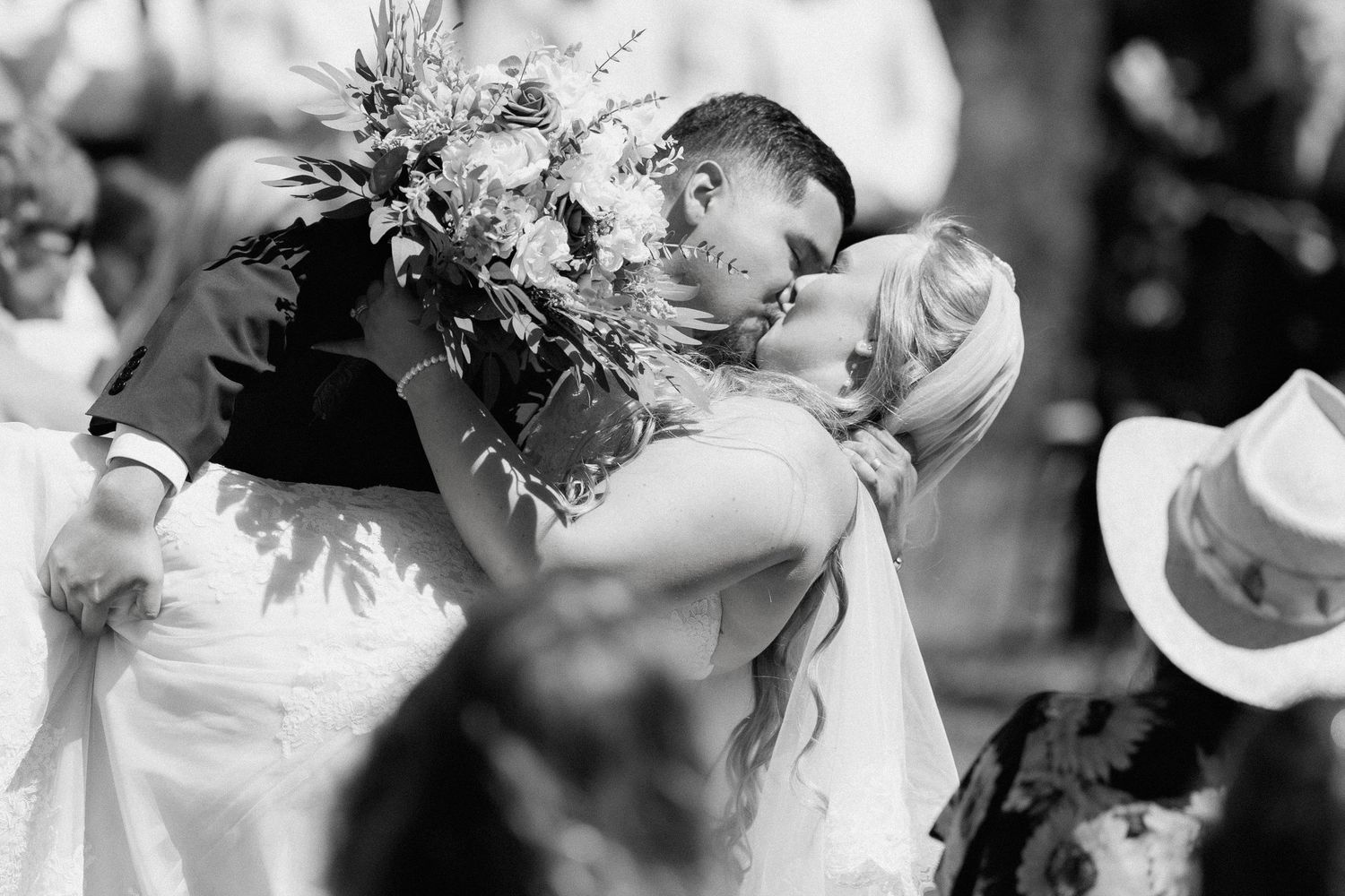 Bride and groom share a romantic first kiss surrounded by wedding guests in black and white photo.
