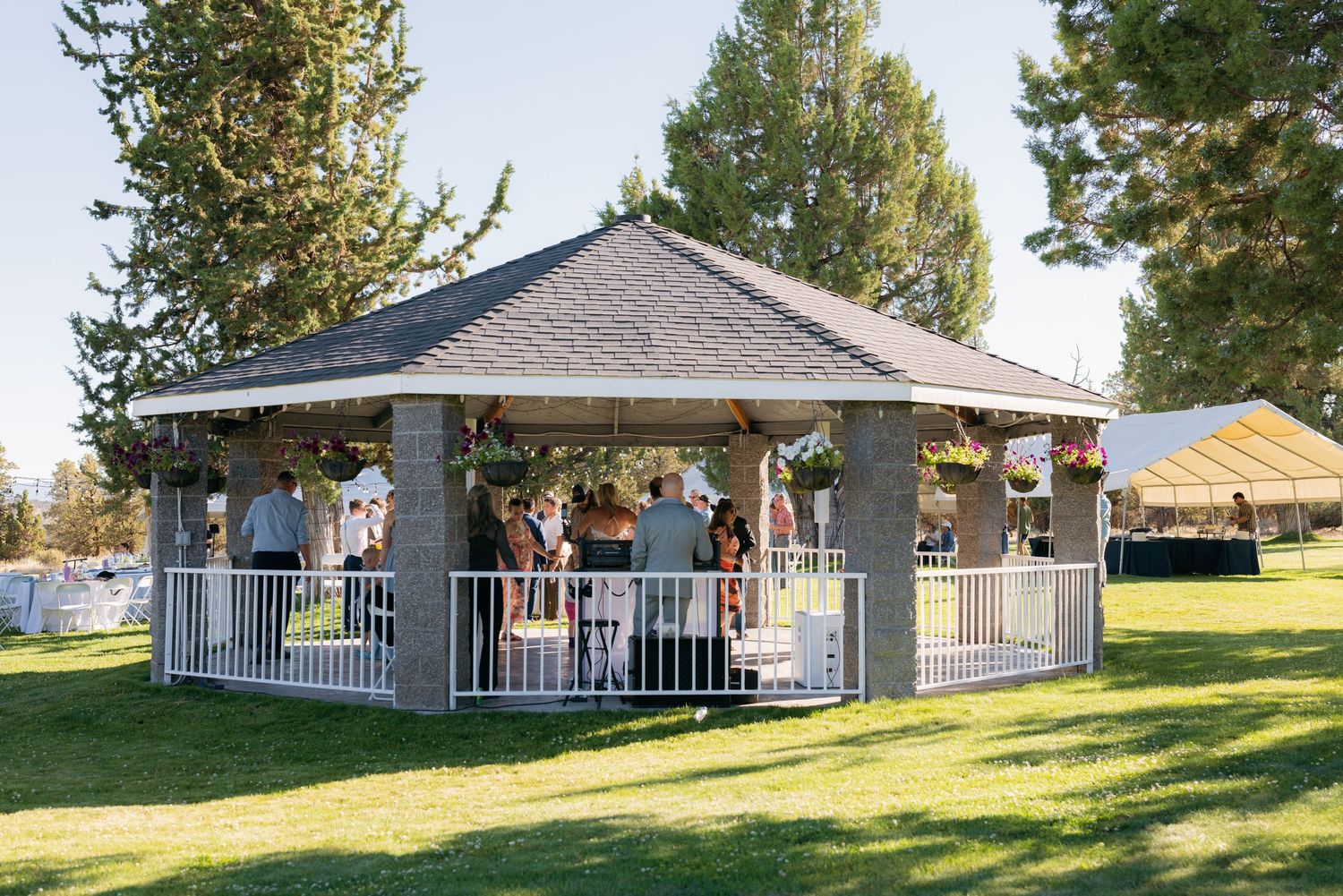 Guests gather inside a gazebo decorated with flowers during an outdoor wedding reception.