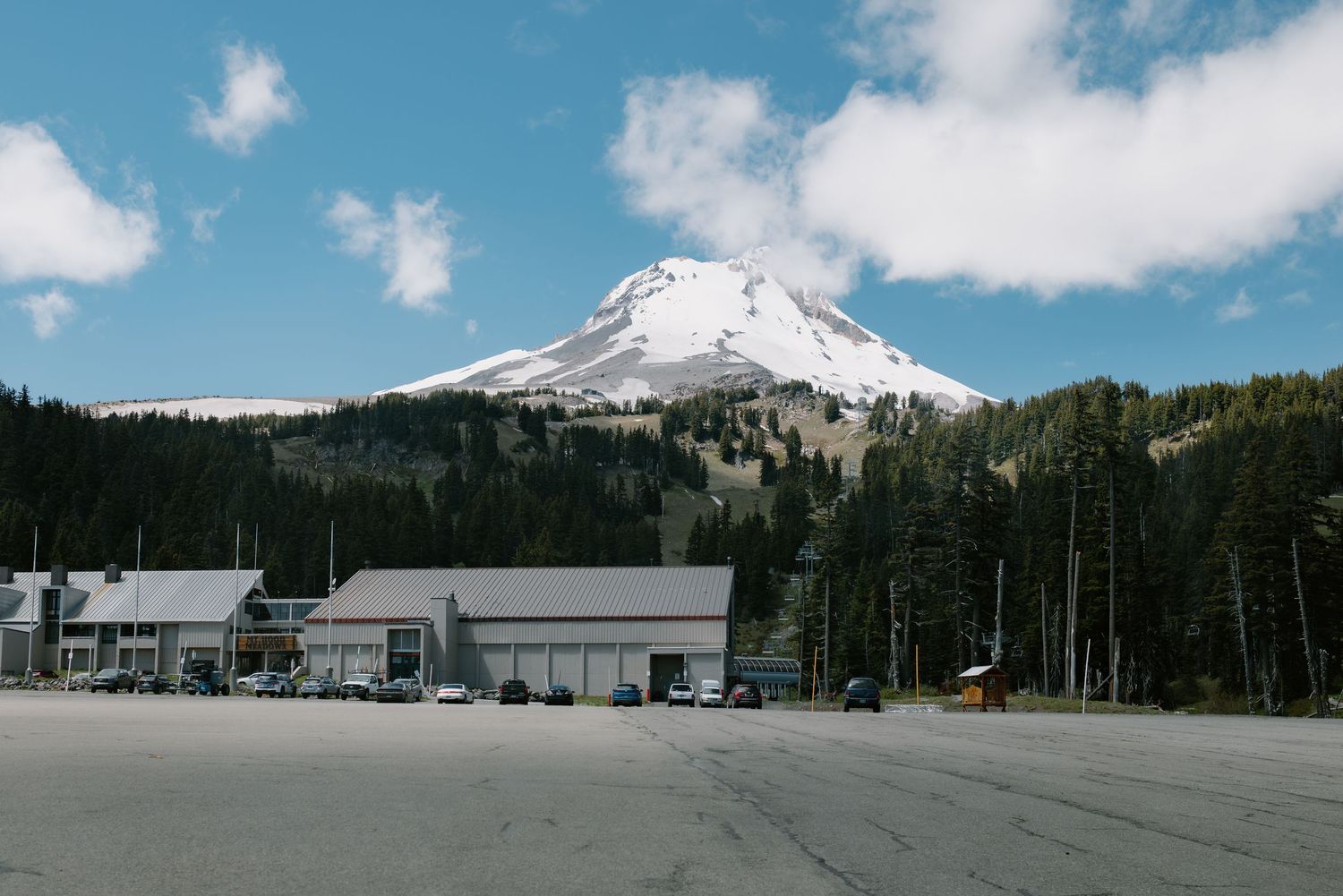 Snow-capped Mount Hood towers over a parking lot and buildings surrounded by evergreen forests under a partly cloudy sky.