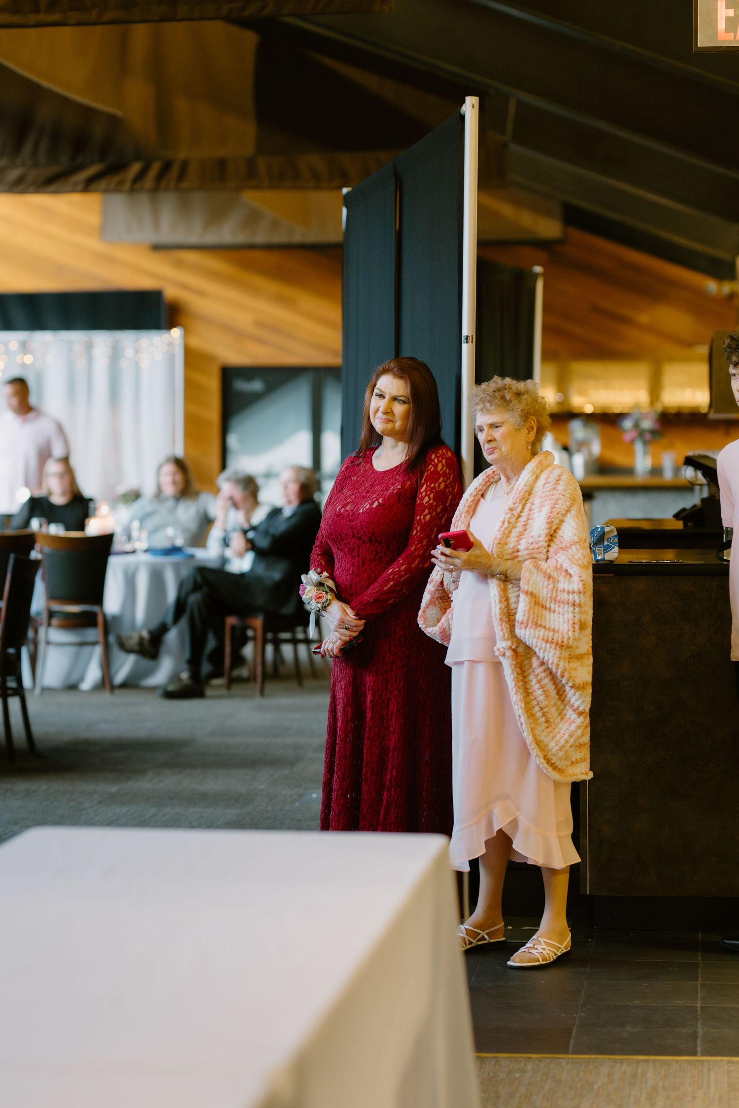Two women in formal attire stand together at an elegant indoor wedding venue with guests seated in the background.