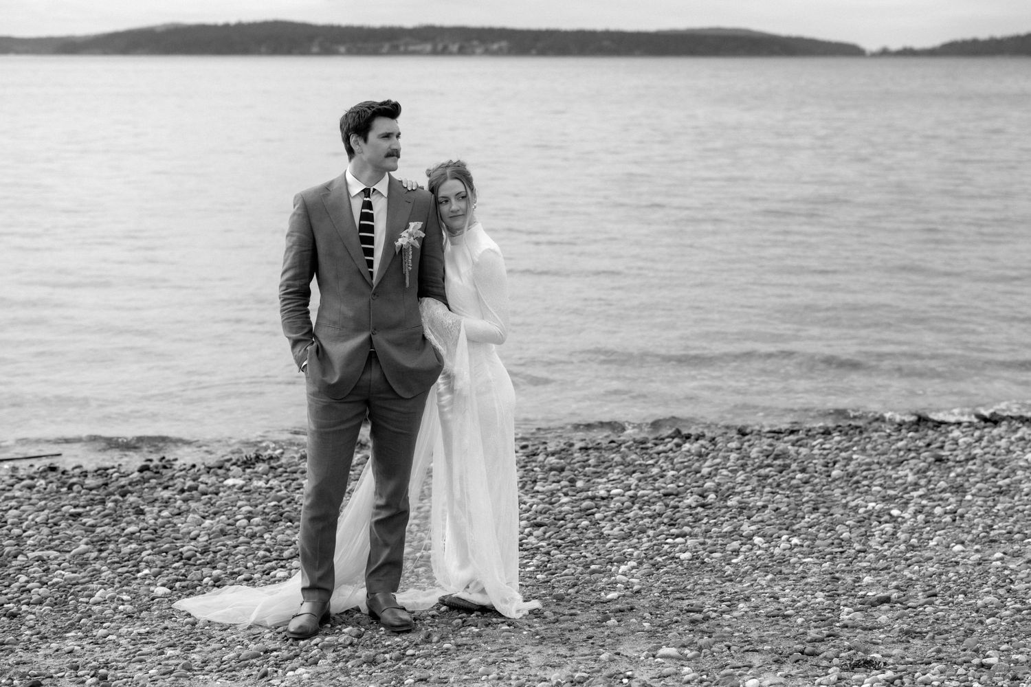 Black and white photo of a bride and groom standing on a rocky beach shore with water in the background.