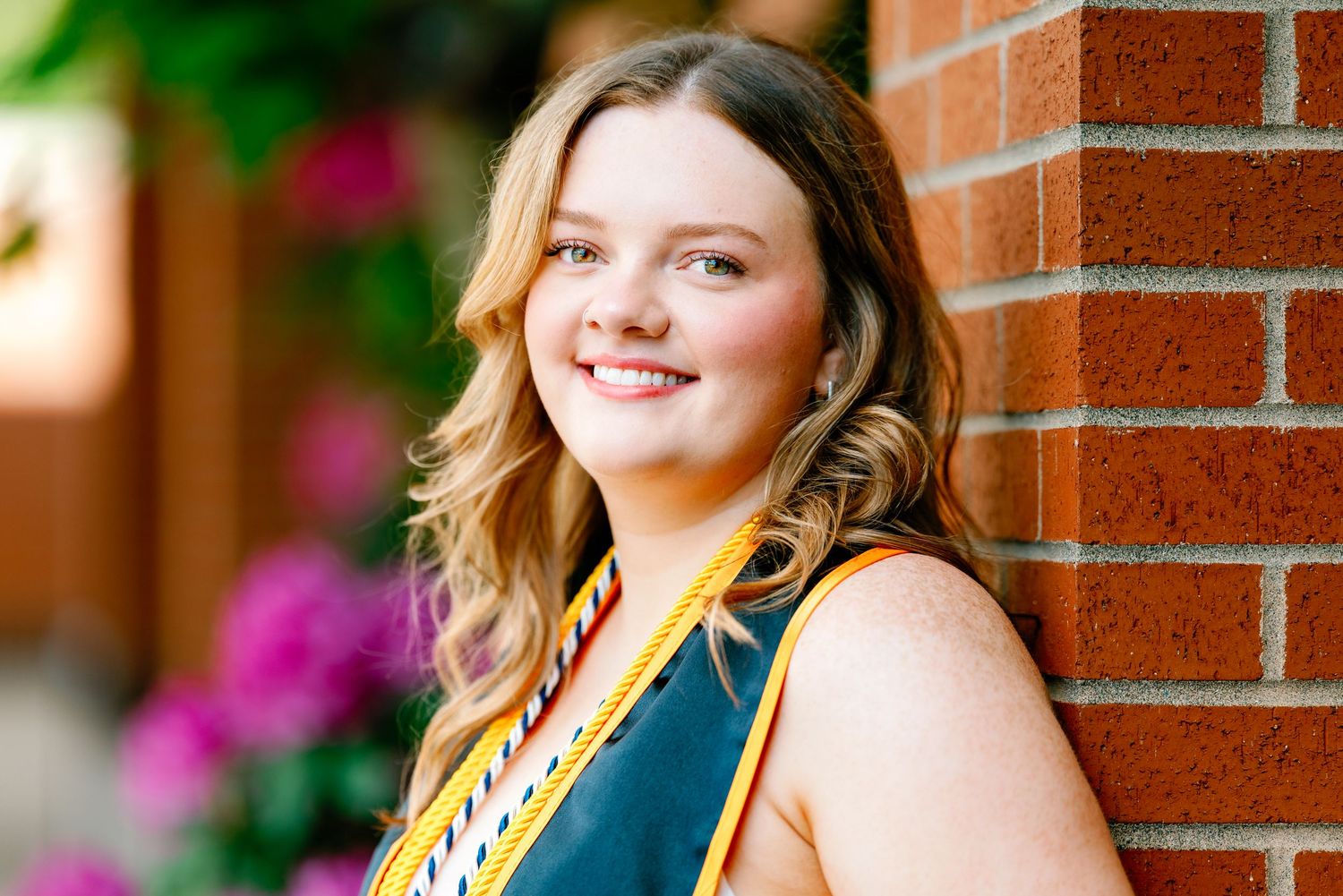 Smiling young woman in graduation attire leaning against a brick wall with colorful flowers in background.