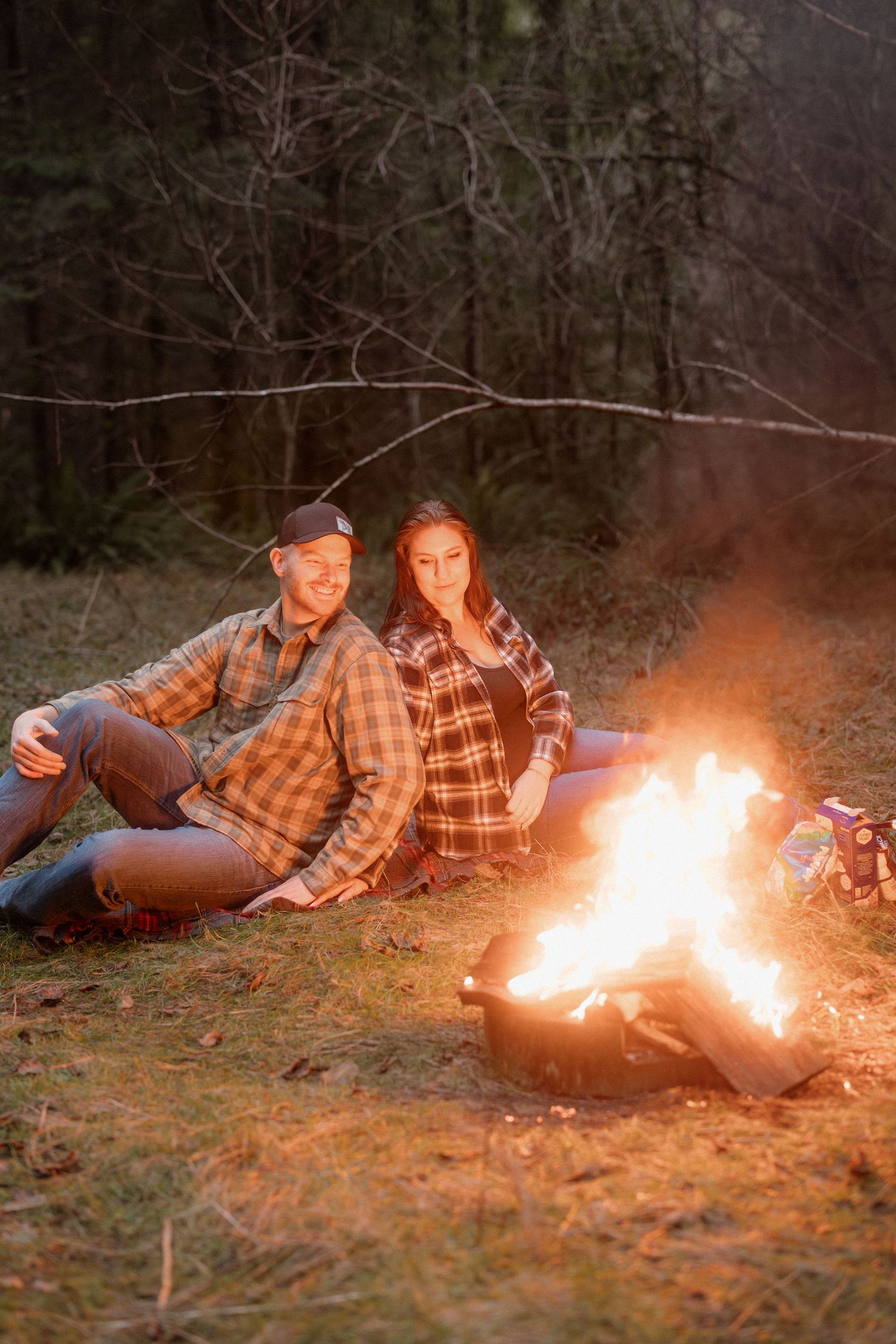 Two people in plaid shirts lying on the ground near a bright campfire in a dark wooded area at night.