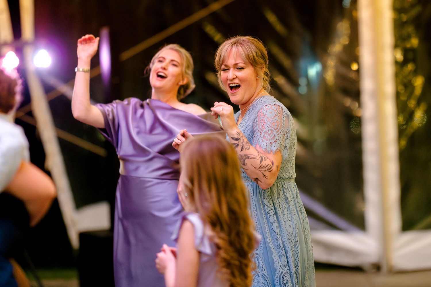 Joyful wedding guests dancing and celebrating on a dance floor illuminated by purple lighting at an evening reception.