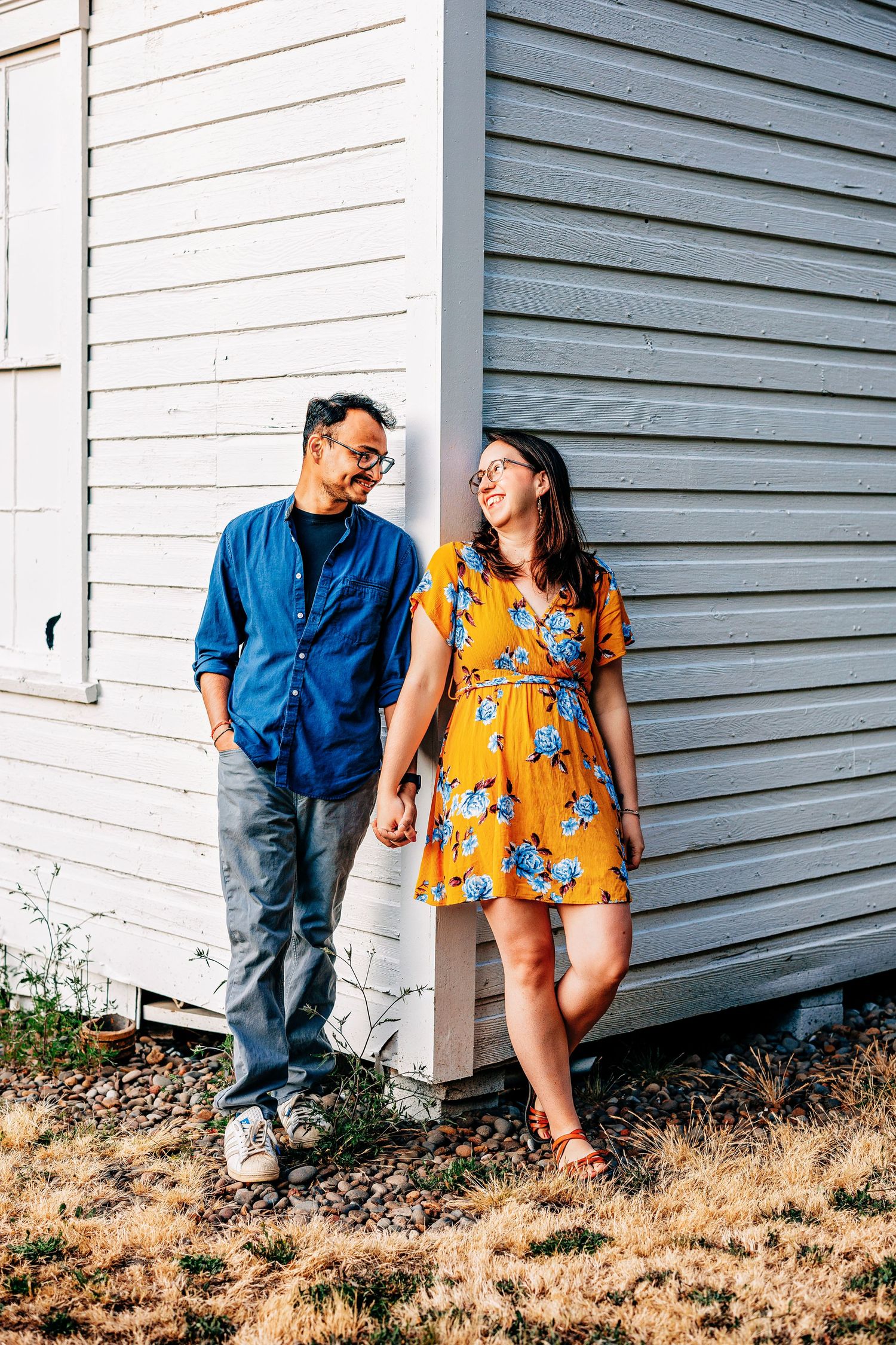 Couple in casual outfits leaning against white building wall, with person in blue shirt and yellow floral dress.
