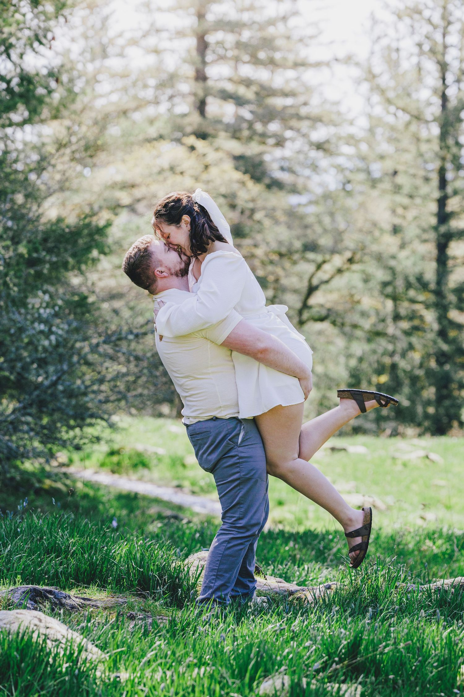 A couple in white embraces playfully on a grassy hillside surrounded by evergreen trees.