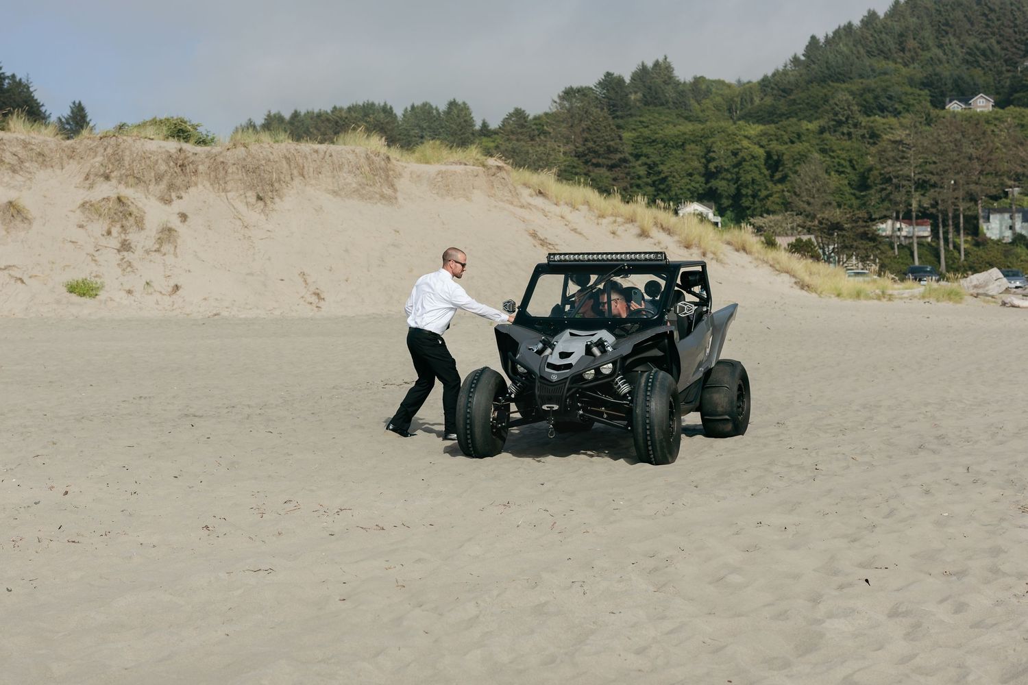 Person pushes dune buggy on sandy beach with forest in background.