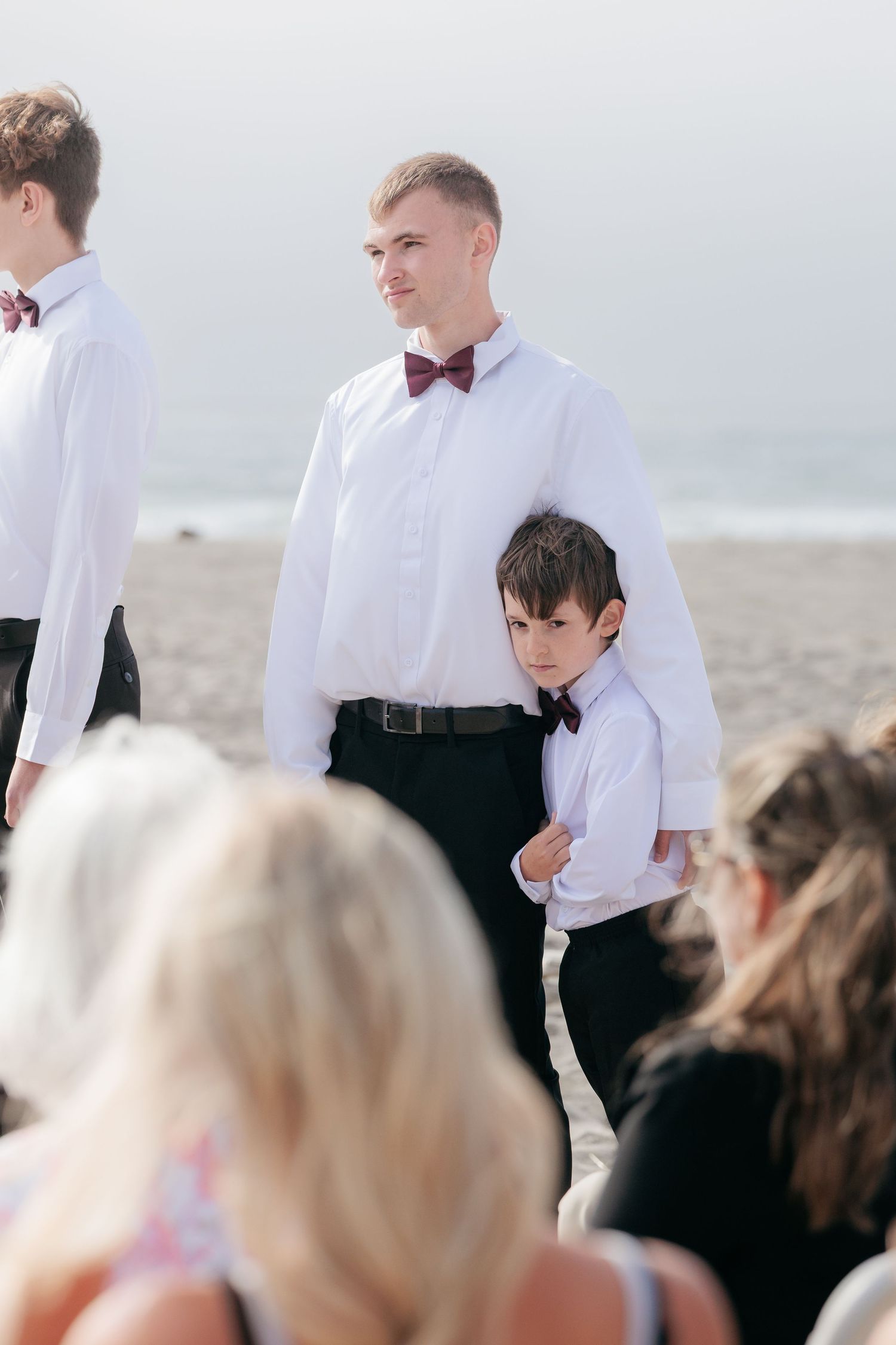 A formal beach wedding scene with attendees in white shirts and bow ties against an ocean backdrop.