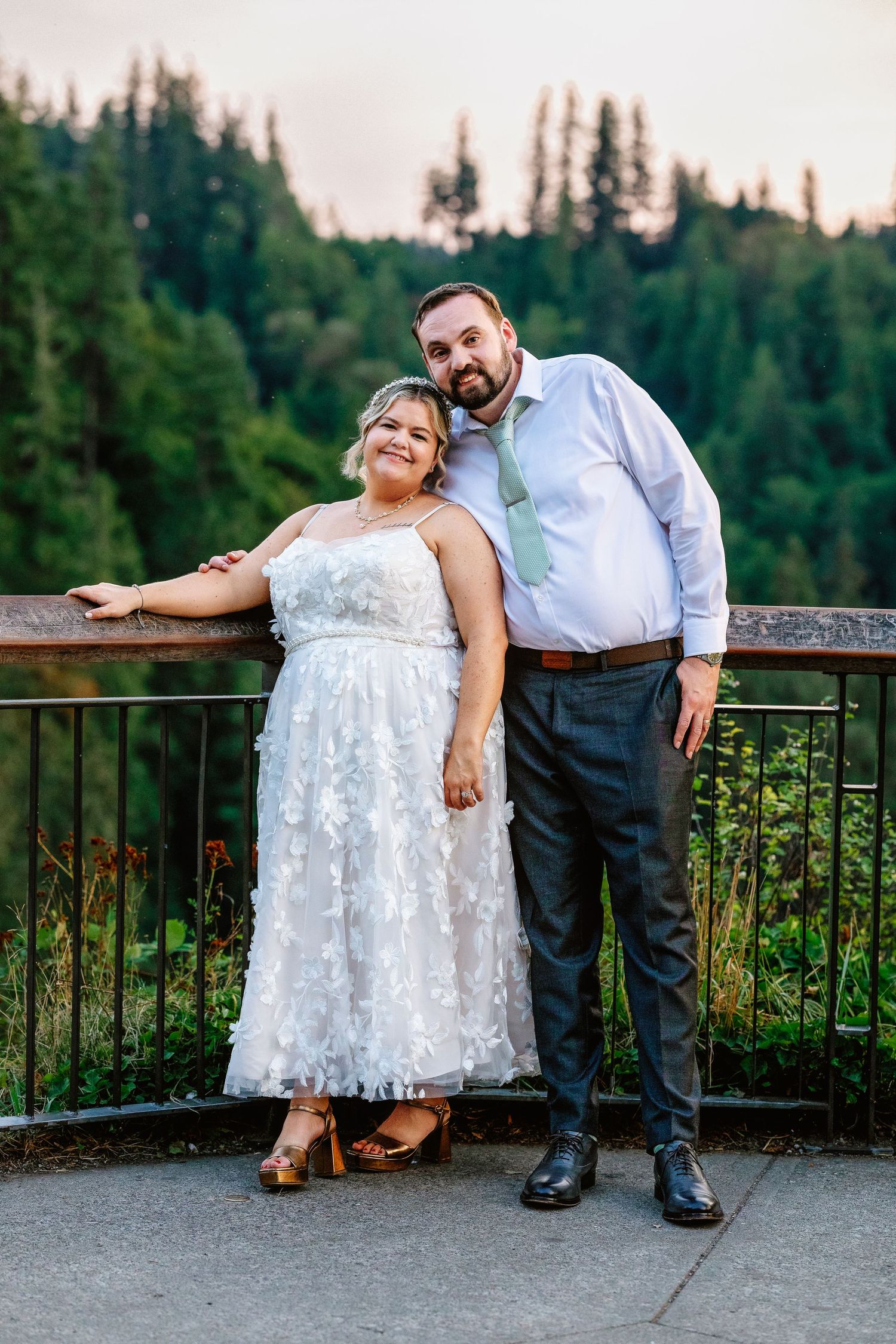 Couple poses together against scenic mountain overlook with forest backdrop during sunset photo session.