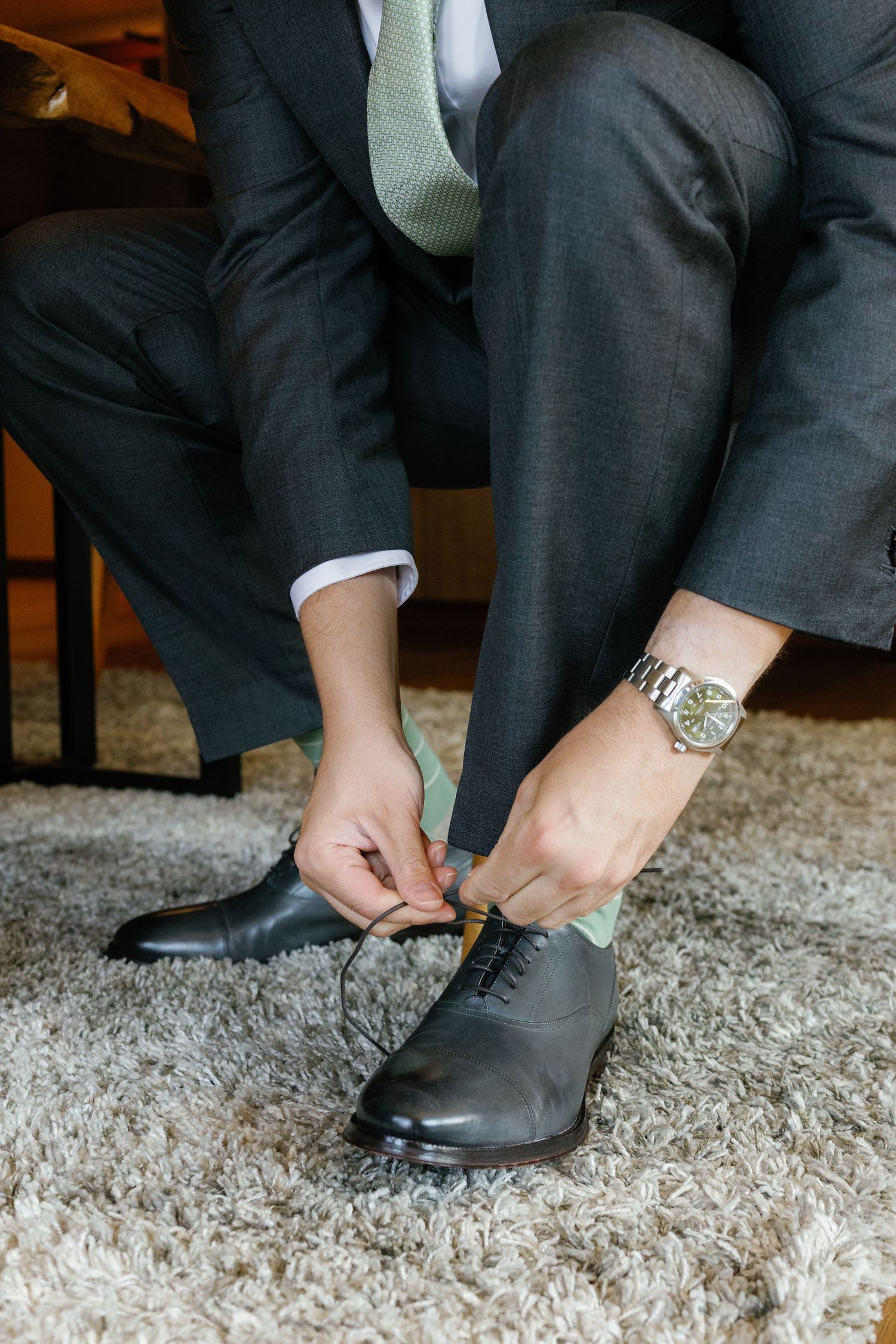 Person putting on formal black dress shoes while sitting on plush carpet.