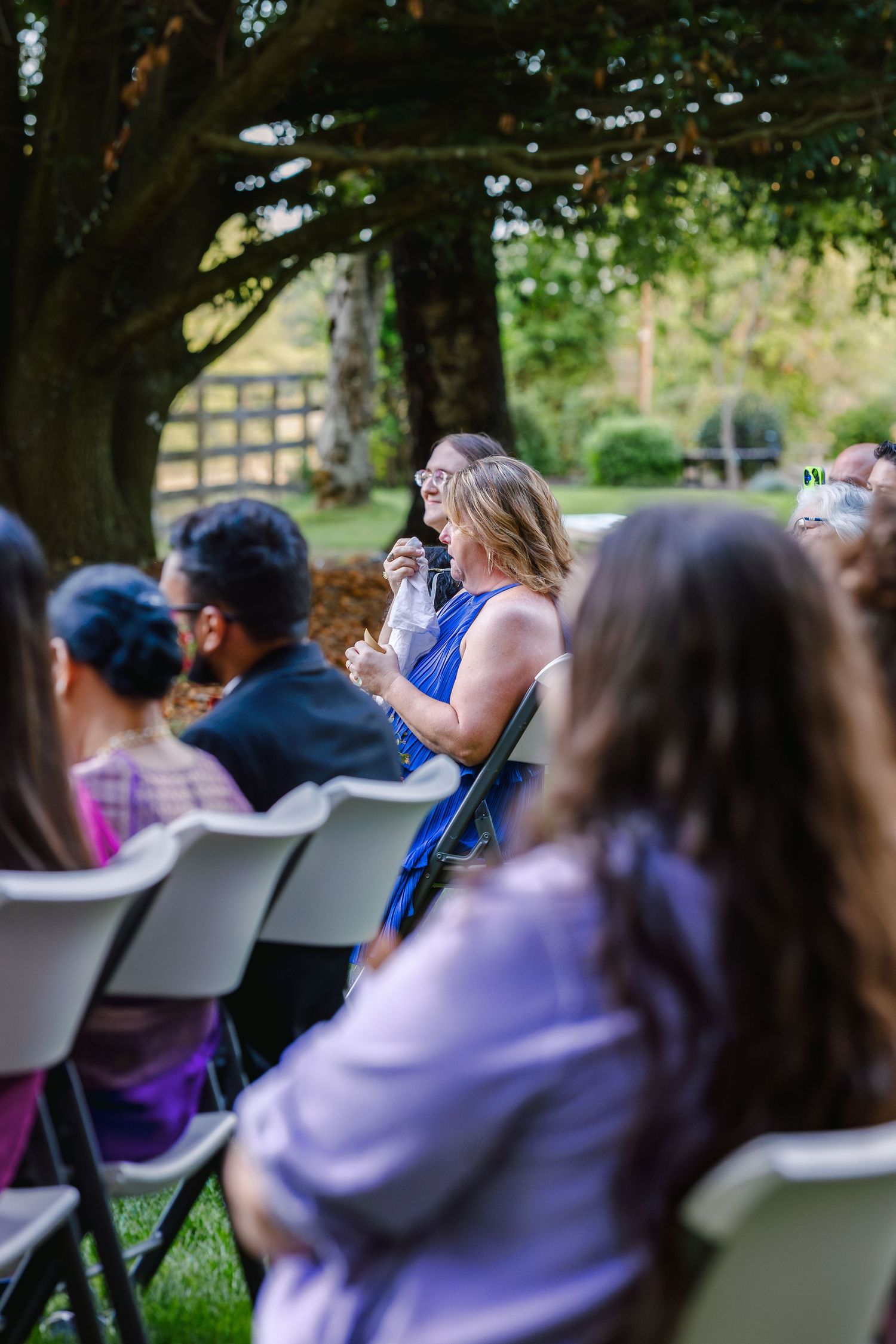 Guests seated in white chairs at an outdoor event beneath shady trees.
