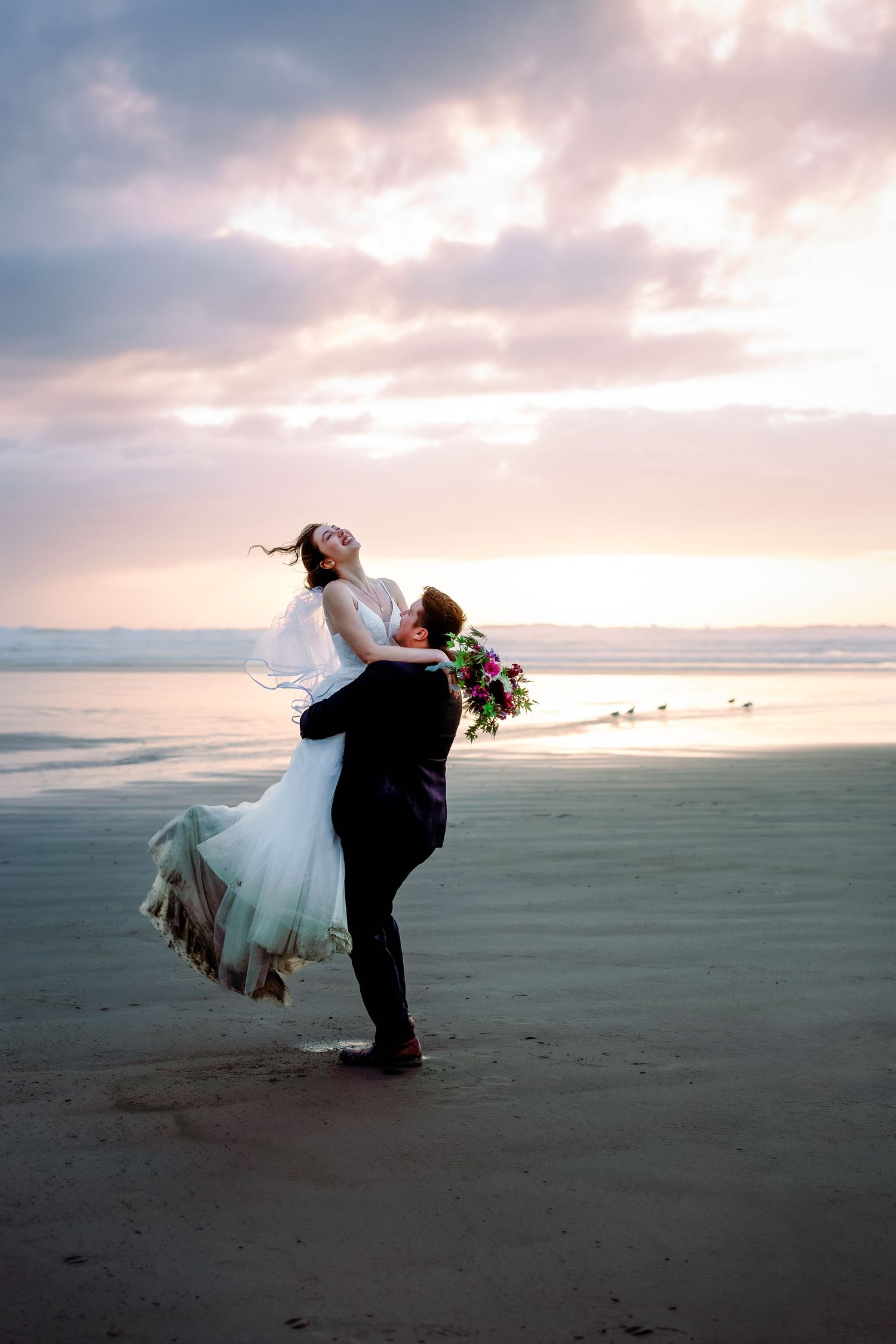 A couple in wedding attire embraces on a beach at sunset, with the groom lifting the bride as her dress flows in the ocean breeze.
