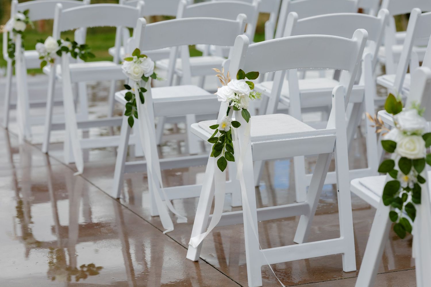 White folding chairs decorated with floral arrangements line up for an elegant outdoor wedding ceremony.