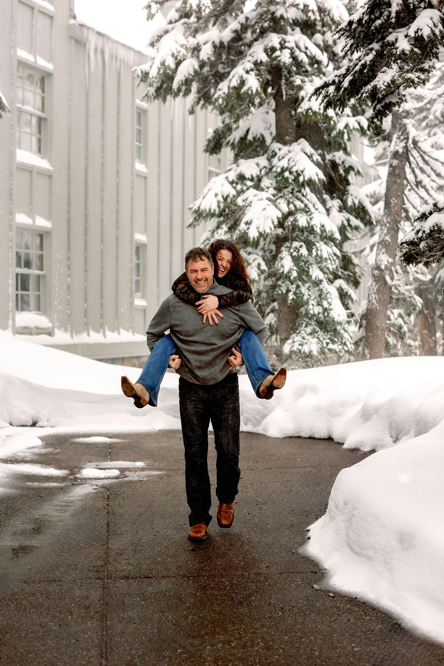 A playful couple frolics in the snow, with one person giving a piggyback ride to another against a snowy backdrop.