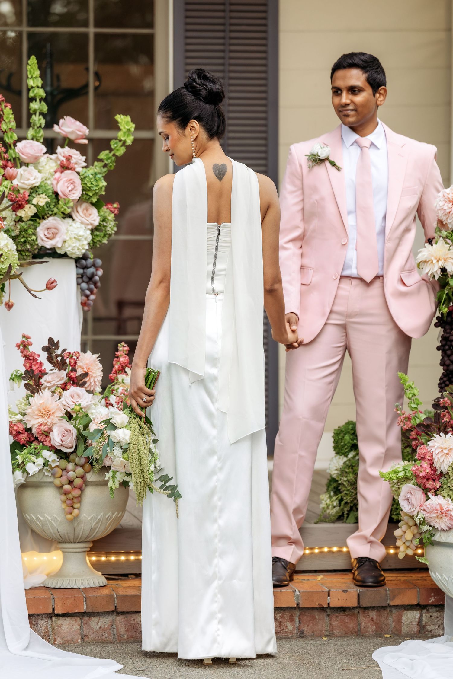 Modern wedding couple in white dress and pink suit pose by floral arrangements on brick steps.