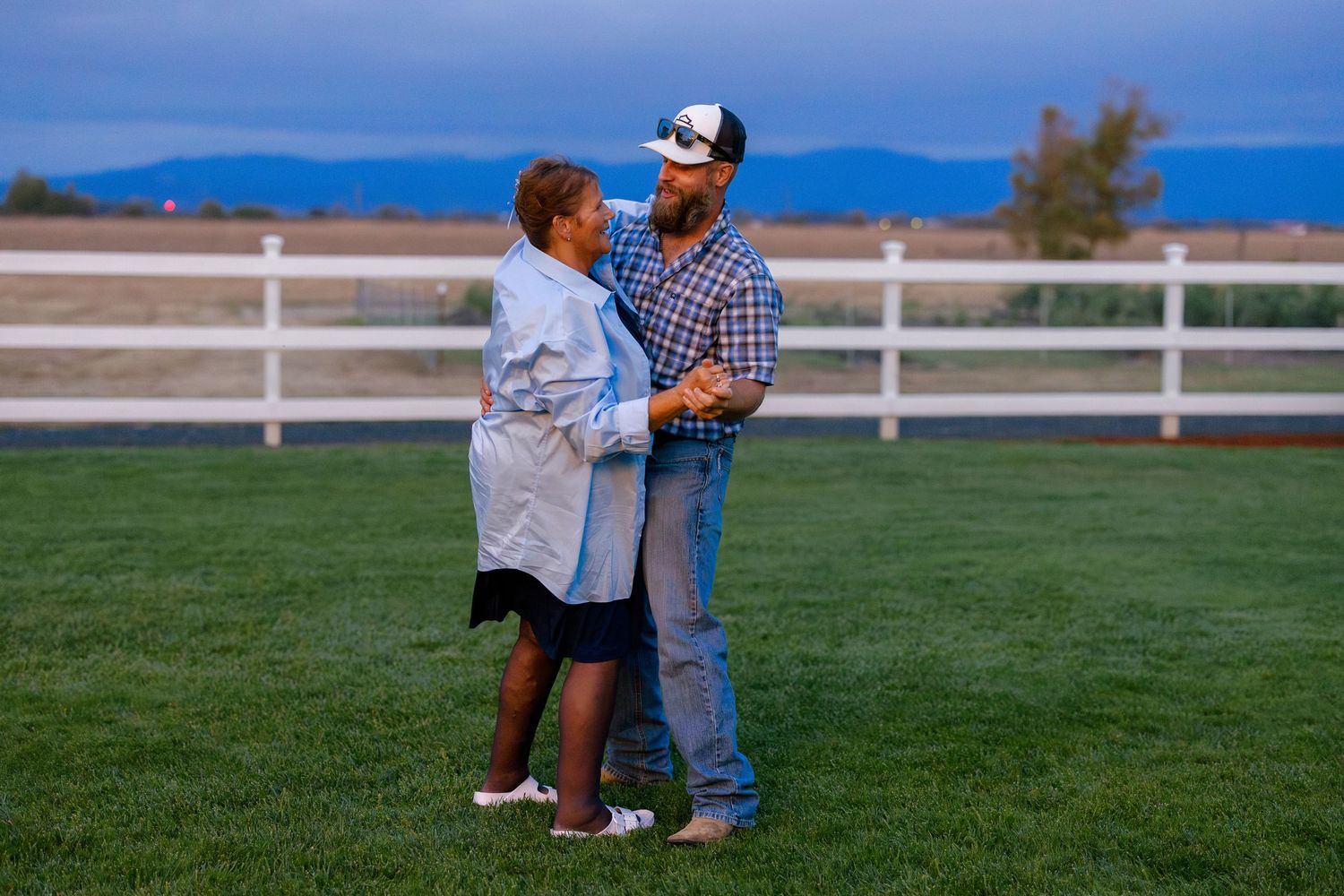 A couple shares an intimate moment on a grassy lawn with white fencing and mountains in the background.