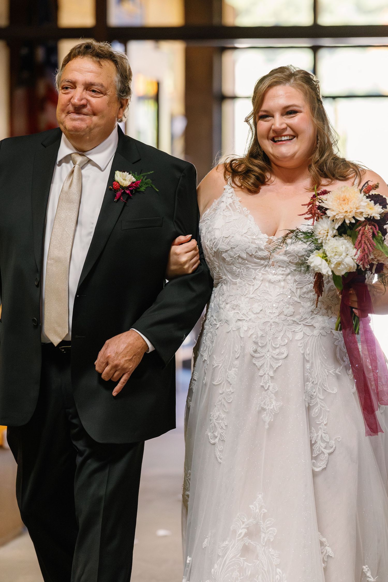Wedding party walks down a sunlit hallway with floral bouquets and formal attire.