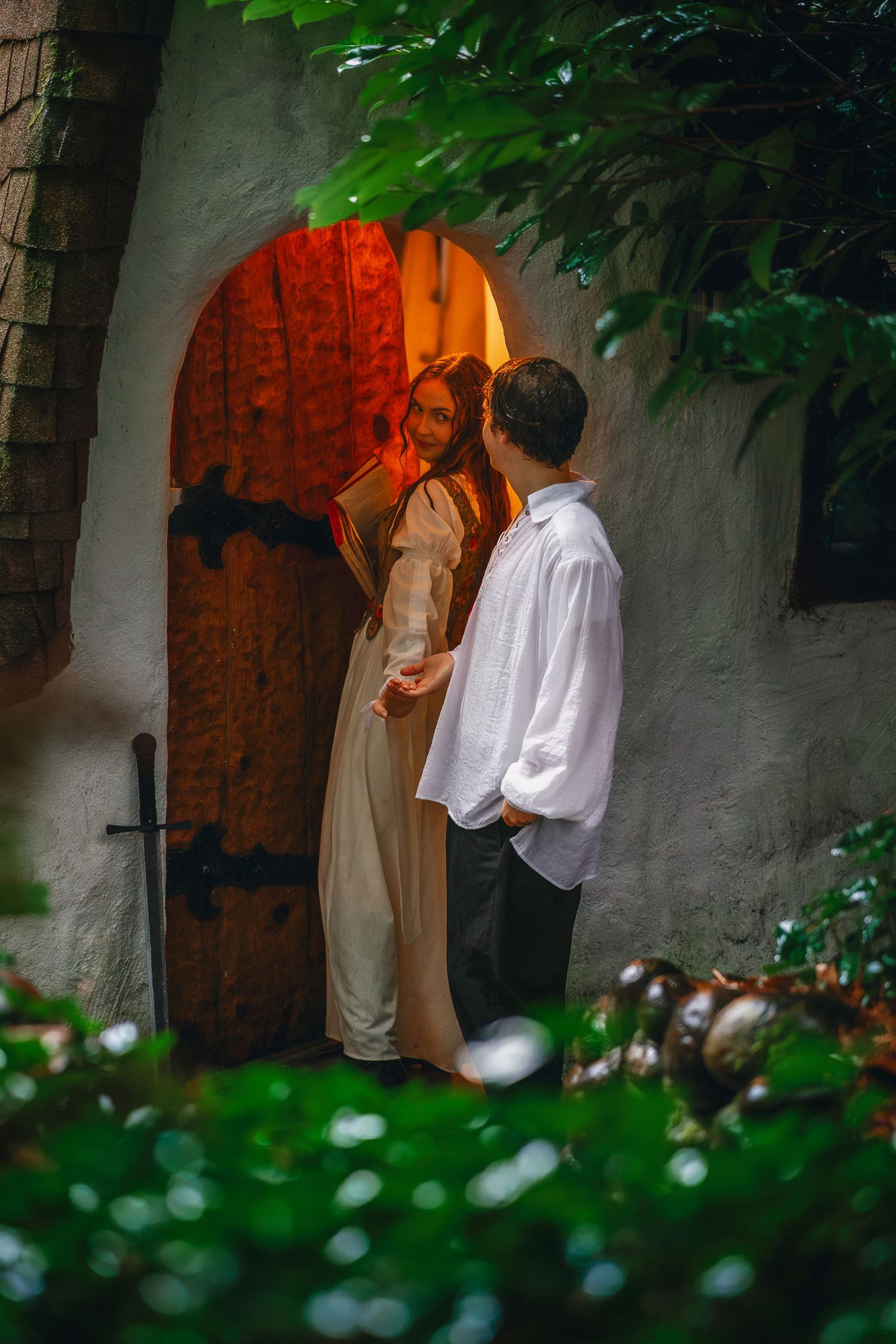 Two figures share an intimate moment by an illuminated red doorway surrounded by greenery.