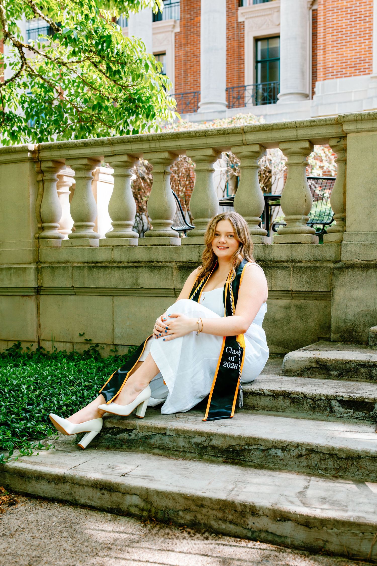 A young woman in graduation regalia with a green and gold stole sits on stone steps outside a brick building.