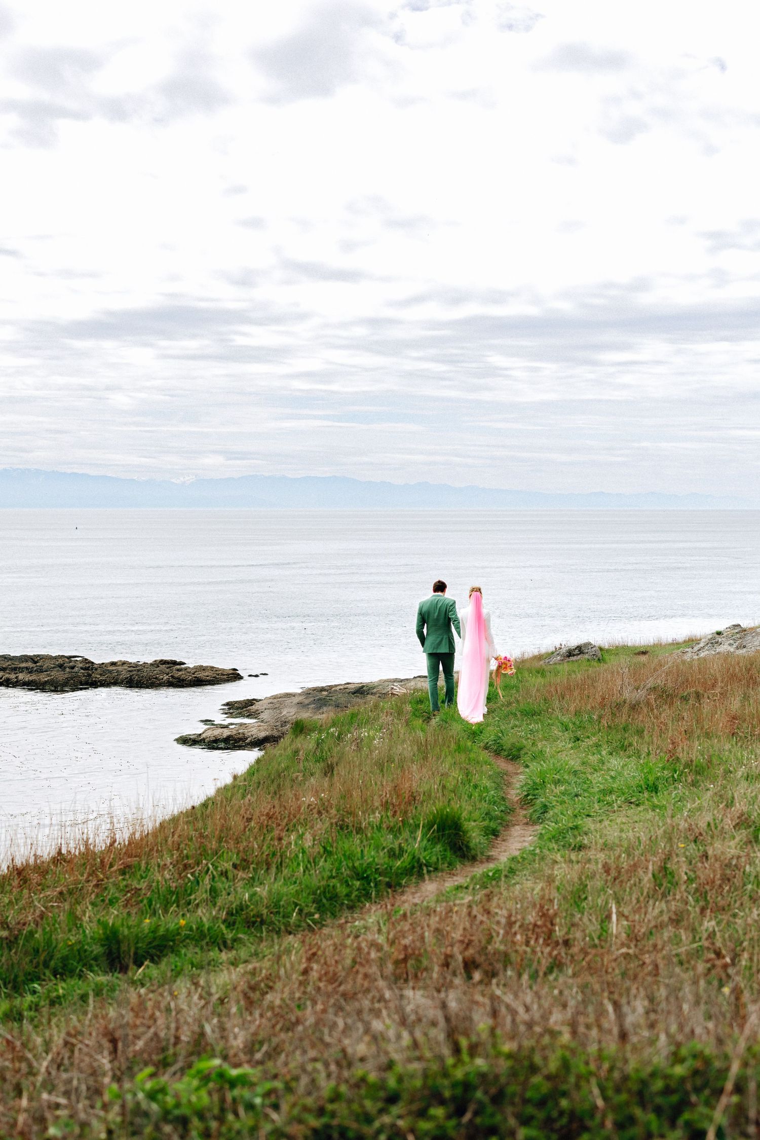 Couple in wedding attire walking along a coastal cliff path, groom in green suit, bride in white dress with pink cape.
