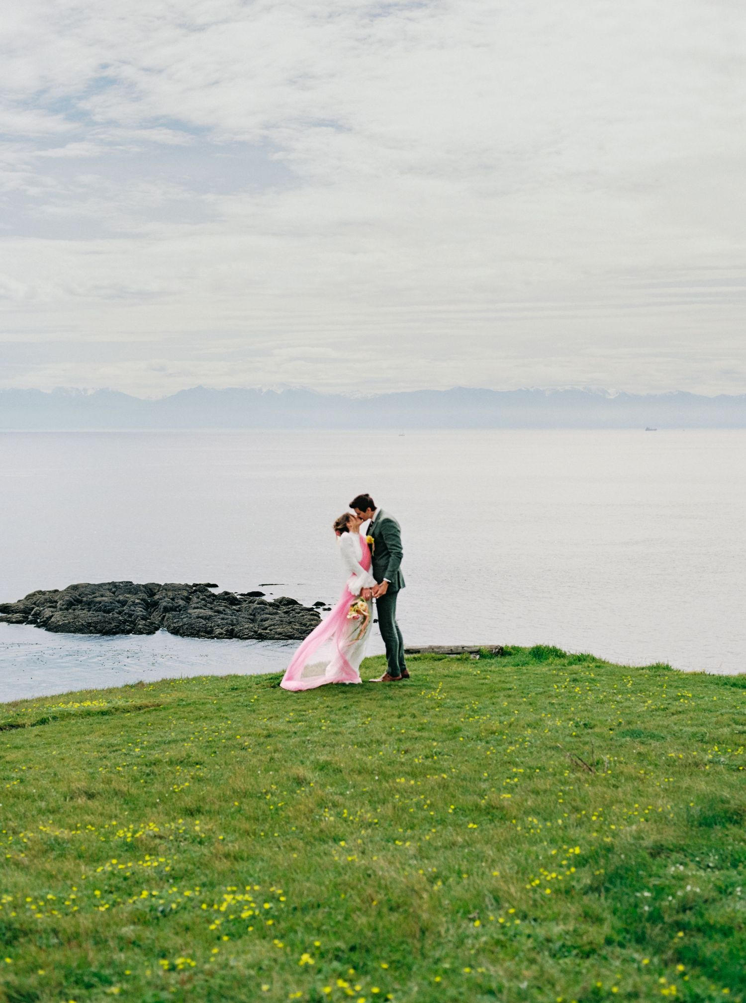 Bride and groom share romantic dip kiss on grassy coastal cliff overlooking serene ocean horizon.