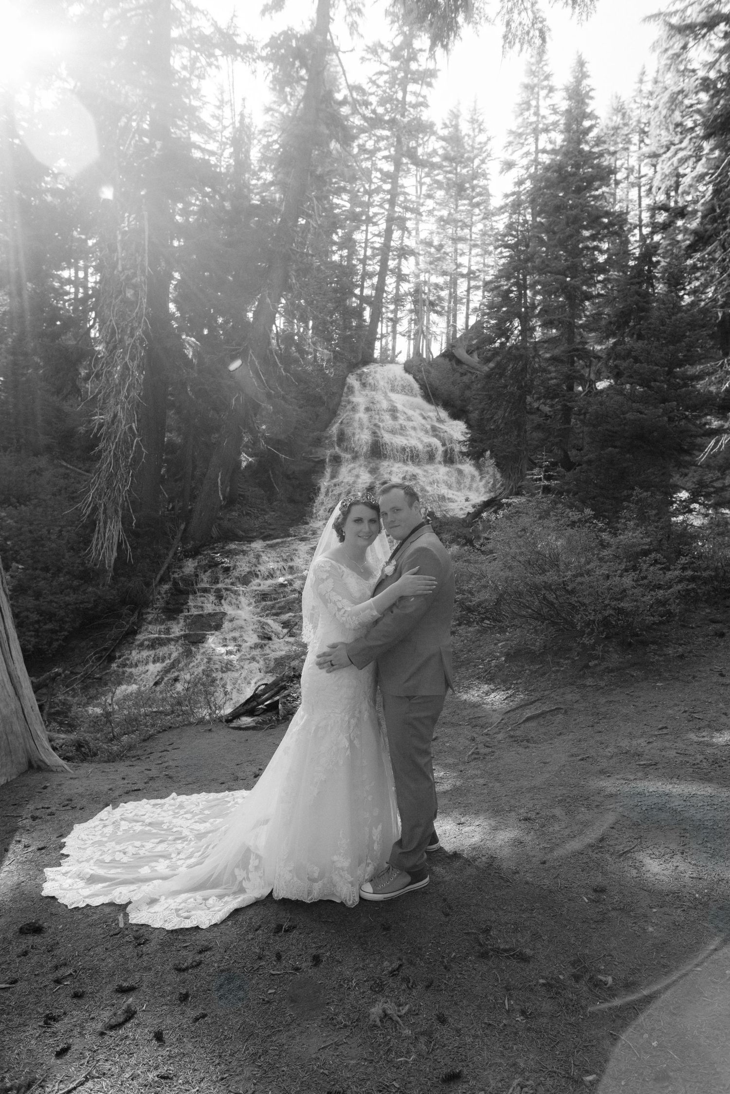 Bride and groom embracing in front of cascading waterfall surrounded by pine trees in black and white.
