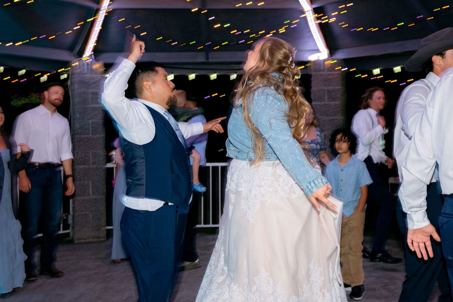 Couple dances joyfully at outdoor wedding reception under string lights surrounded by guests.