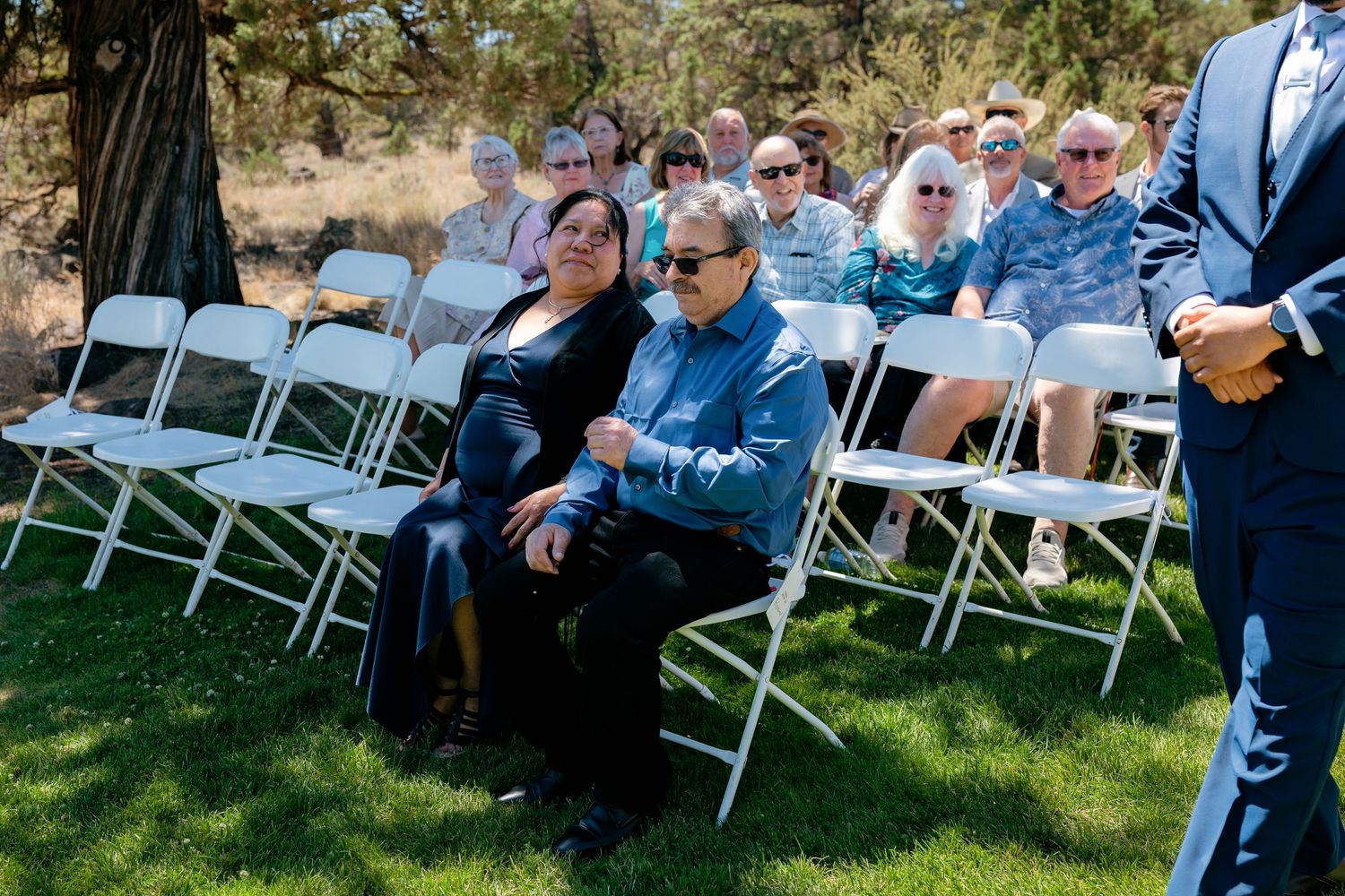 Wedding guests seated in white folding chairs outdoors during a sunny ceremony.