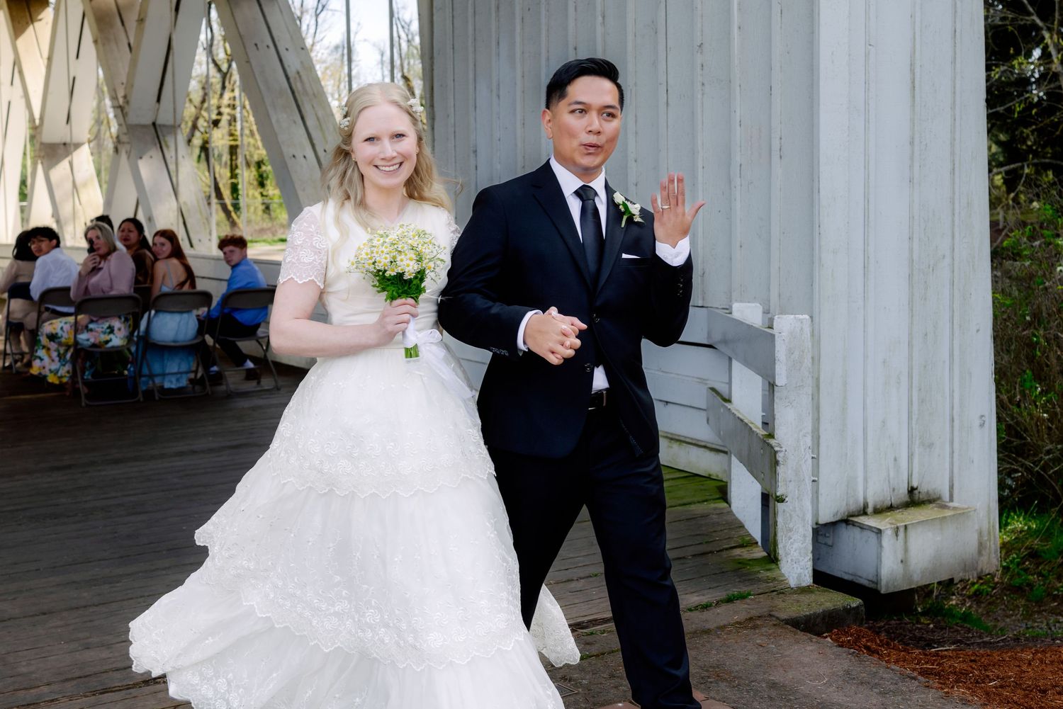 Bride in white gown and groom in black suit walk together waving at outdoor wedding ceremony.