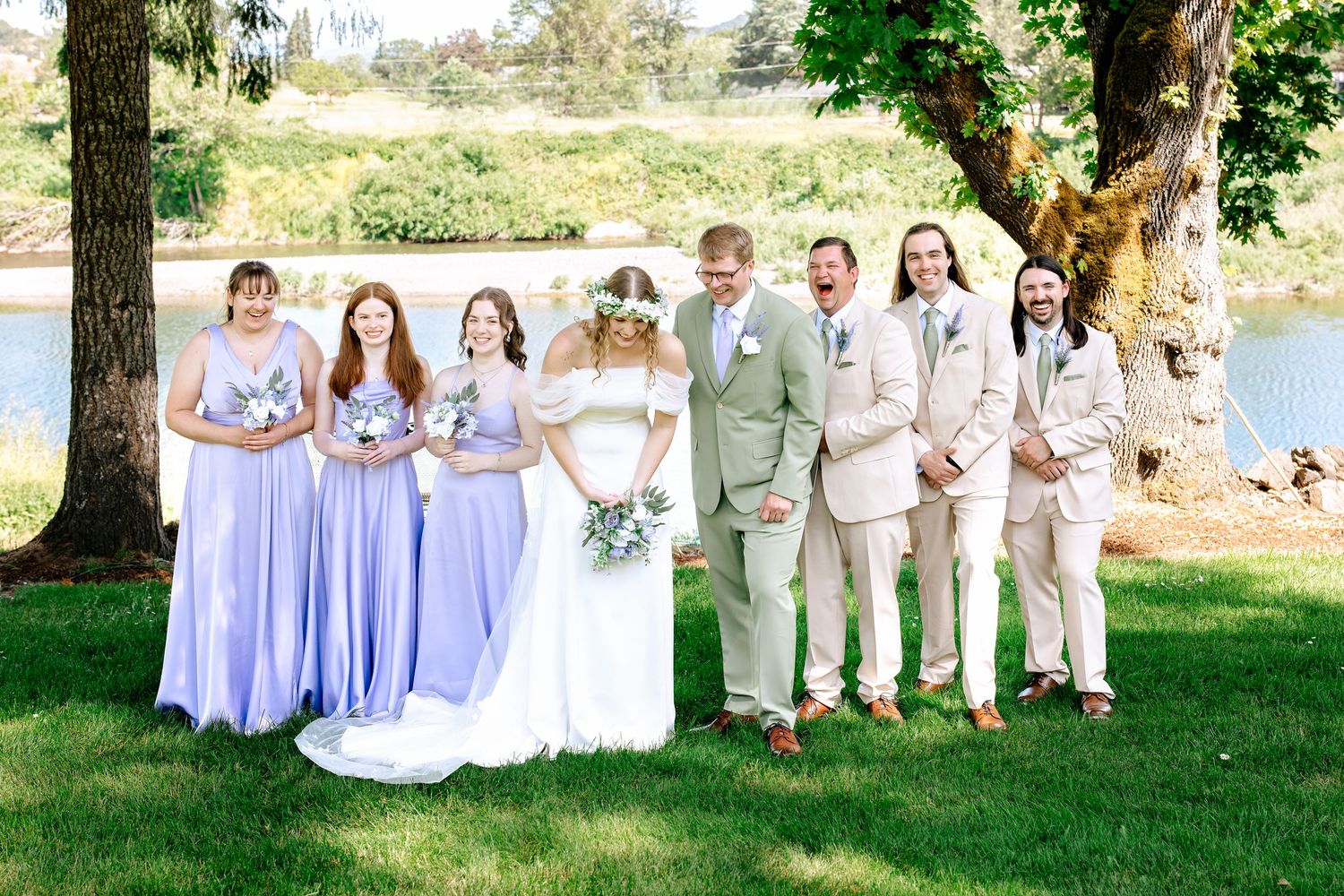 Wedding party poses by a pond, bridesmaids in lavender gowns and groomsmen in tan suits with the happy couple.