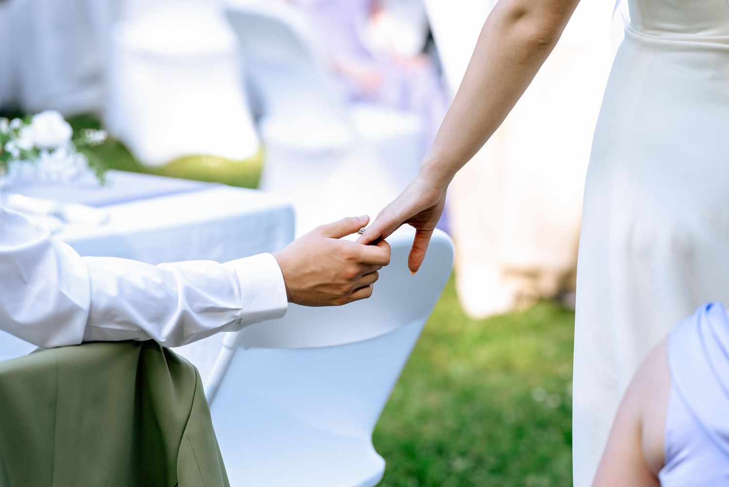 Groom in sage suit gently holding bride's hand in white dress at outdoor wedding reception.