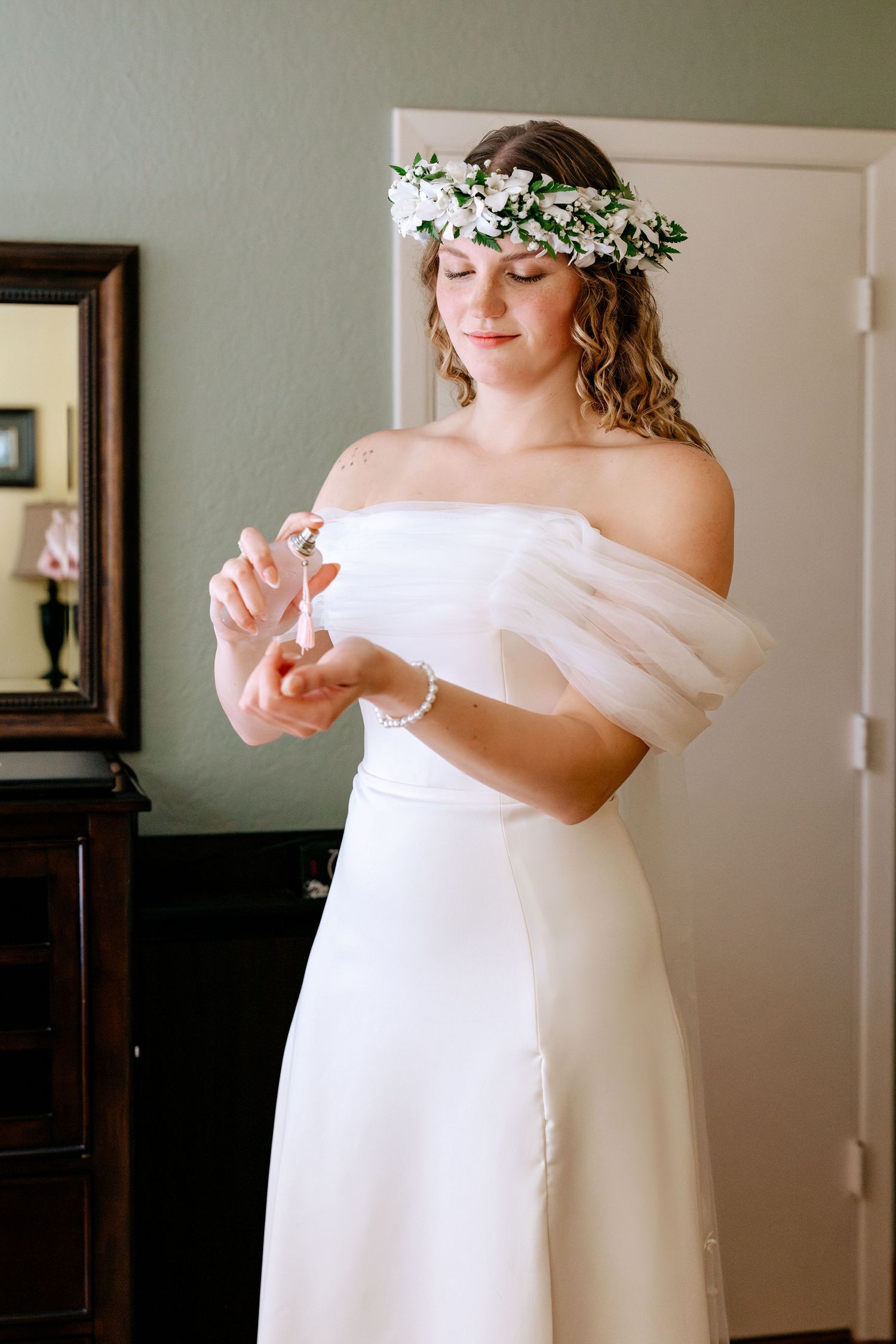 Bride in white off-shoulder gown and floral crown applying perfume while getting ready for her wedding day.