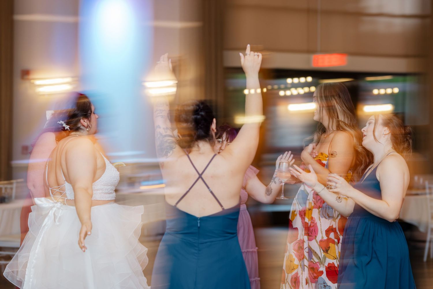 Bride and bridesmaids dancing joyfully at a wedding reception with colorful lights and motion blur.