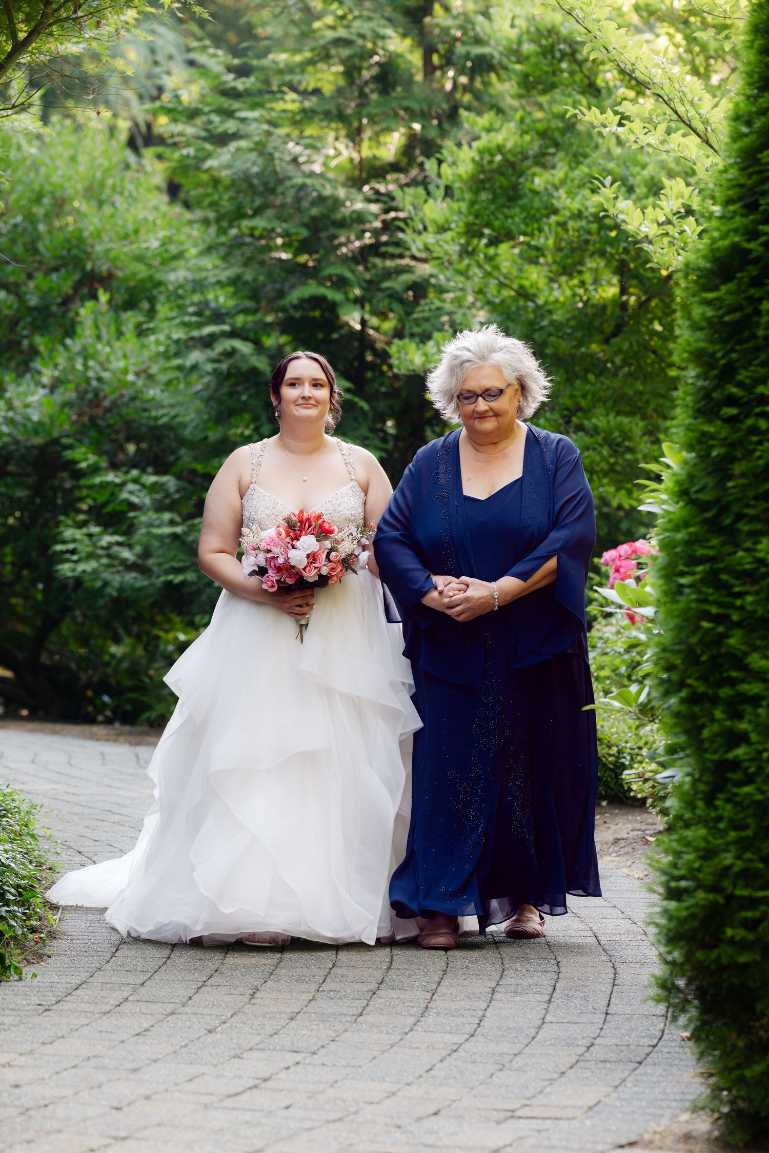 Bride in white layered gown holding pink bouquet walks with older woman in navy dress on garden path.