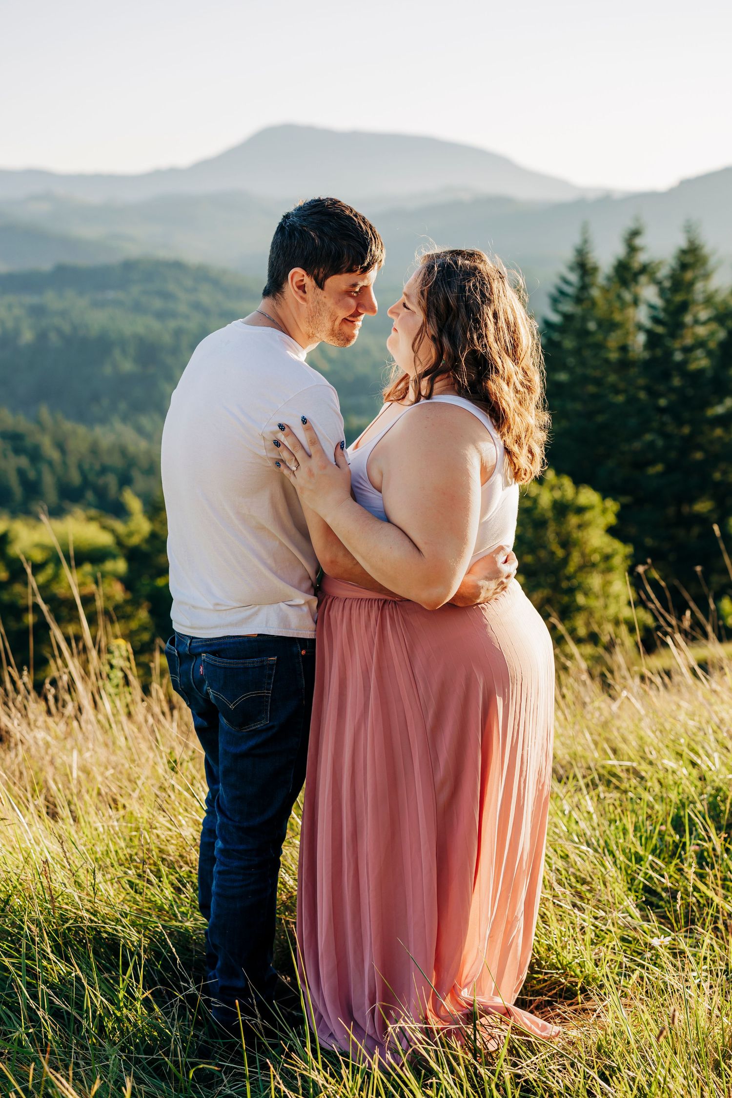 A loving couple embraces on a hilltop meadow at sunset with evergreen forests and mountains in the background.