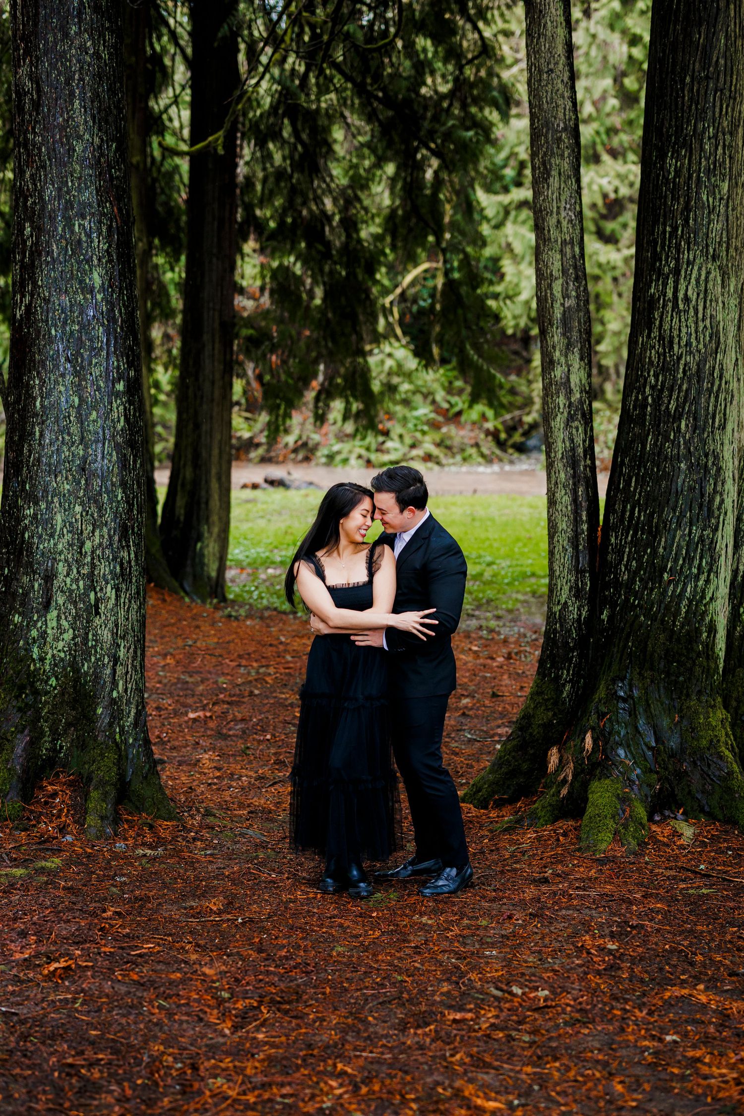 A romantic couple sharing an intimate moment between tall evergreen trees on a forest path covered in fallen needles.