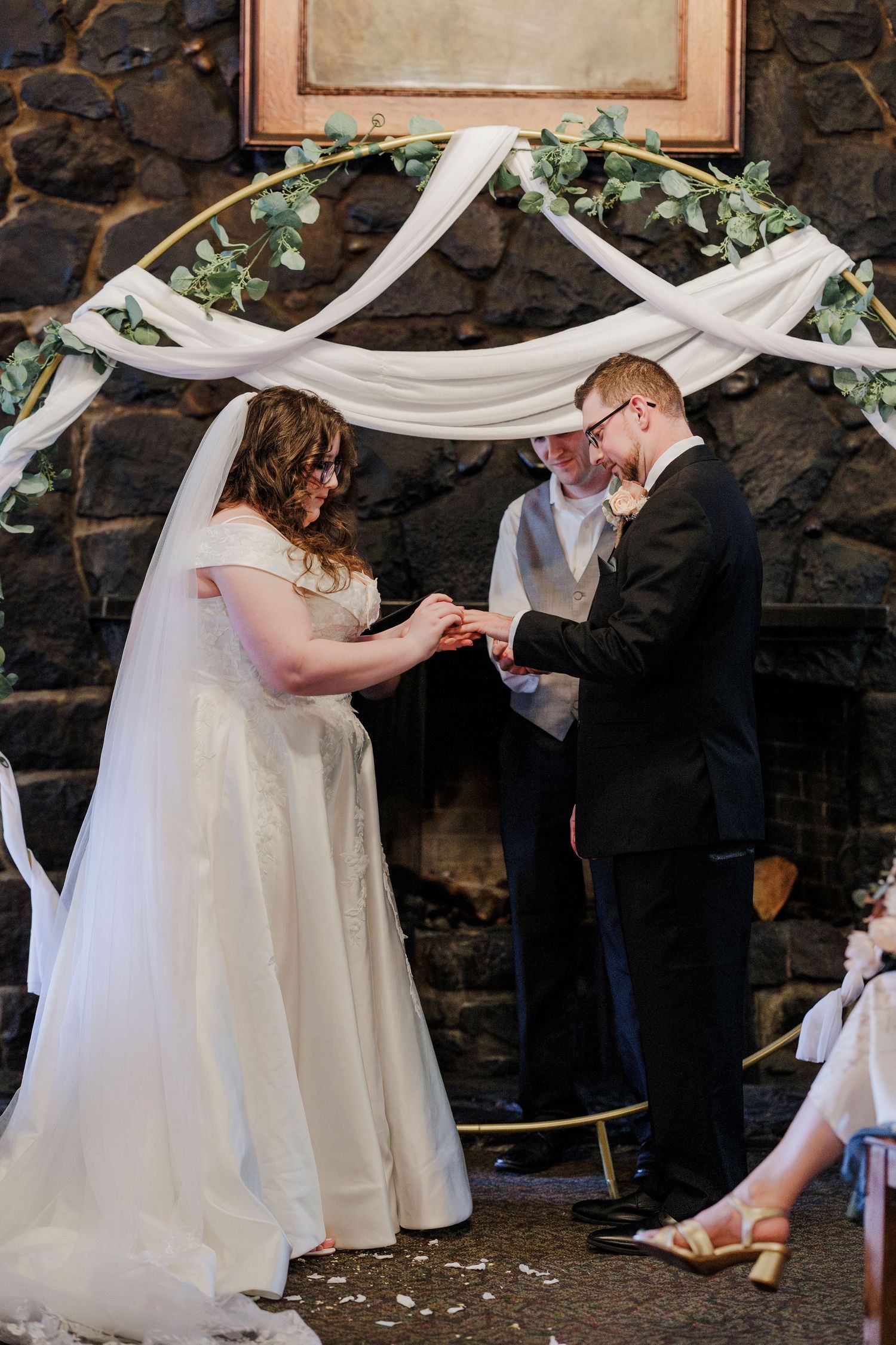 A couple exchanges rings under a white draped arch with greenery during their wedding ceremony.