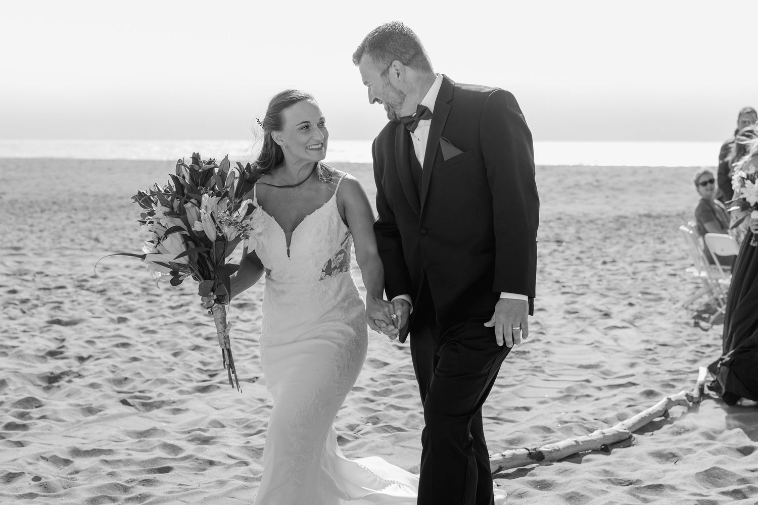Black and white photo of couple walking on beach in formal wedding attire.
