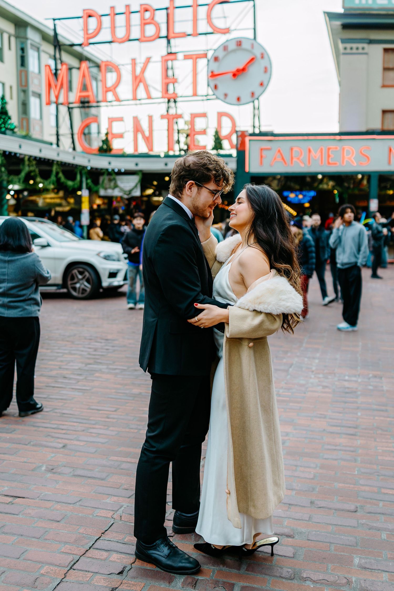 A couple shares a romantic moment outside Pike Place Market in Seattle, with the iconic neon sign visible in the background.