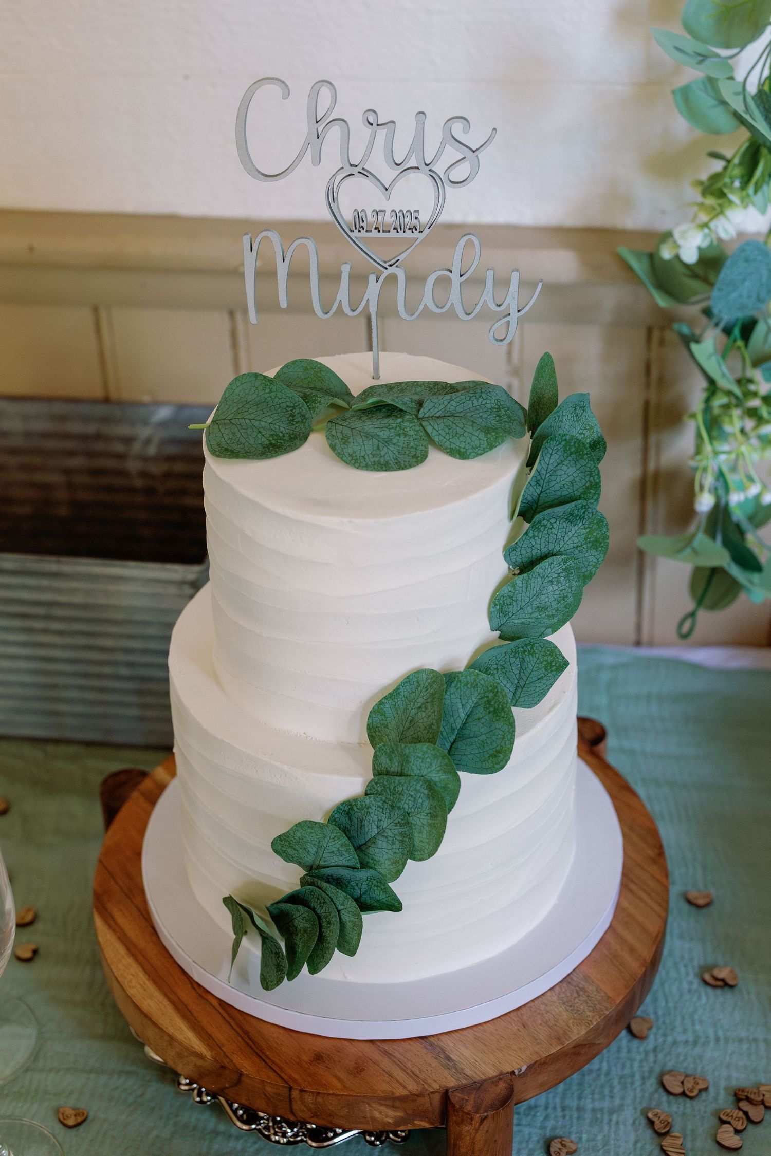 Simple white wedding cake decorated with eucalyptus leaves displayed on a rustic wooden cake stand.